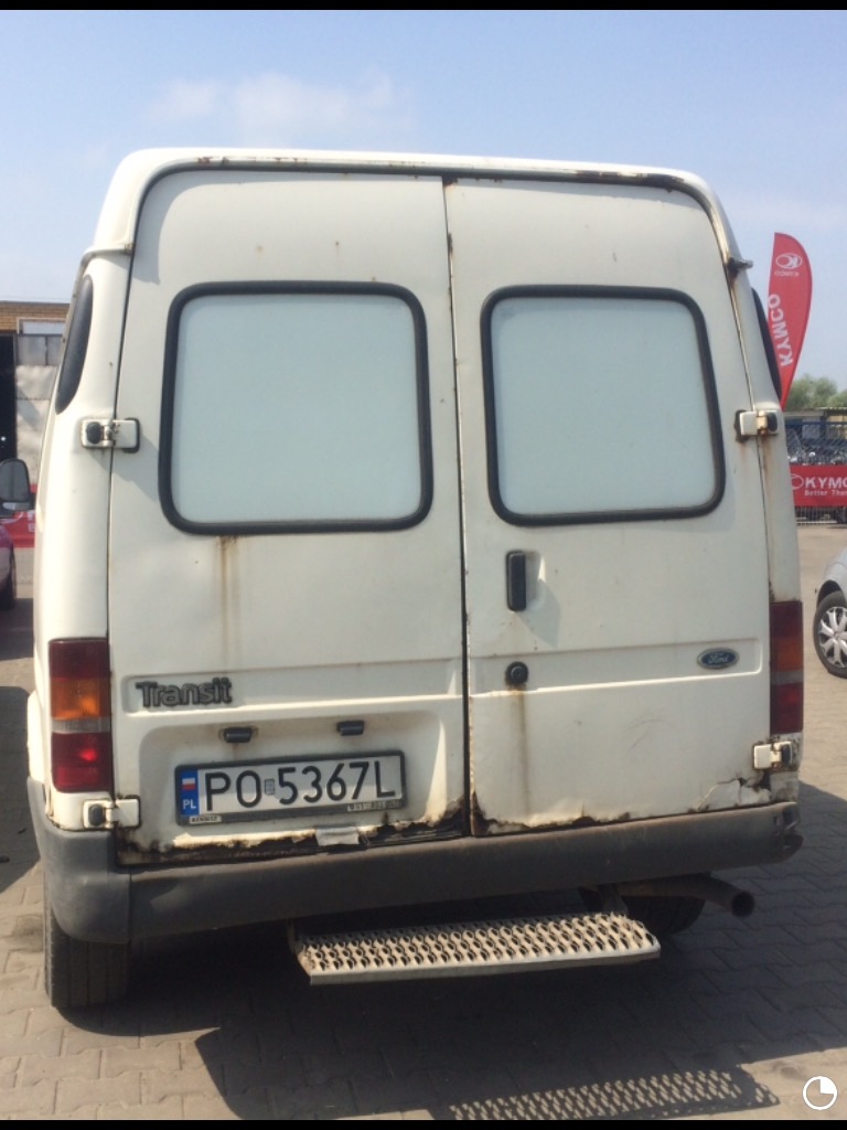 Tylna część białego, zardzewiałego samochodu dostawczego Ford Transit z polską tablicą rejestracyjną, widoczne ślady korozji na karoserii i wokół tablicy rejestracyjnej, metalowy stopień wejściowy.