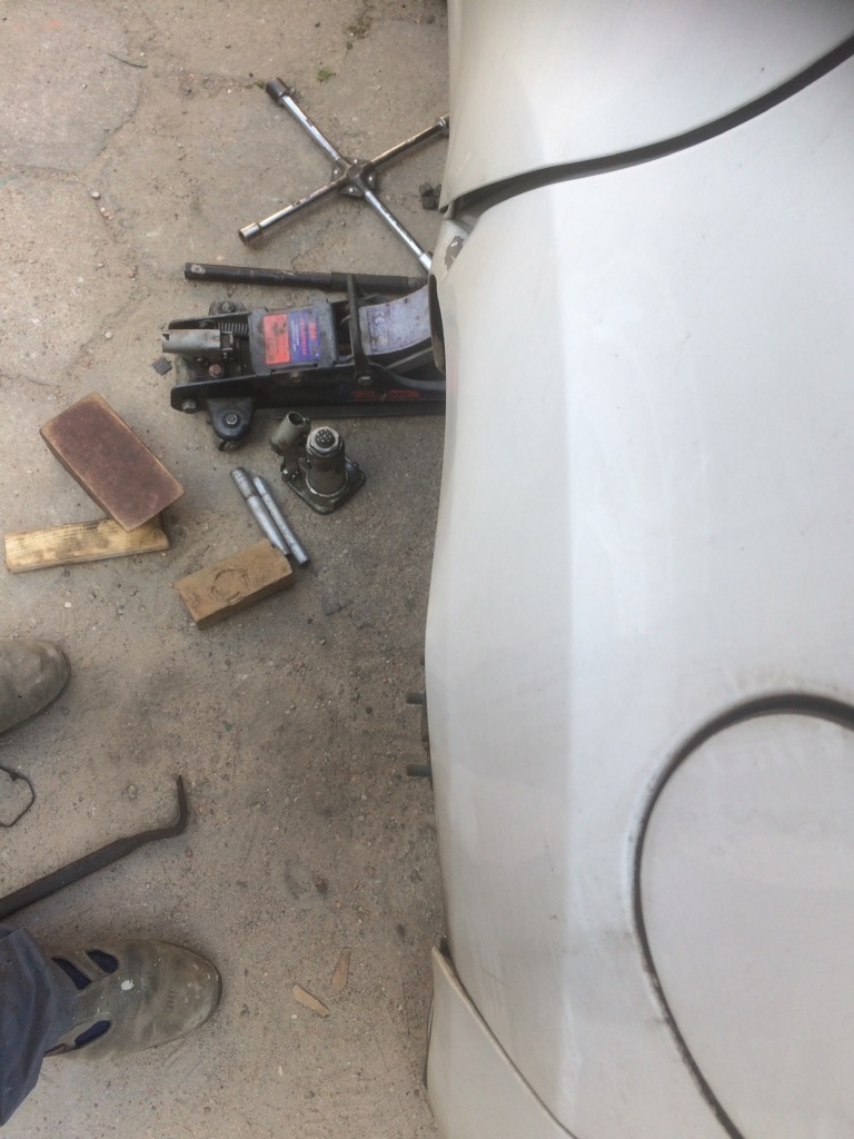 Uszkodzony bok białego samochodu, widoczny podnośnik hydrauliczny, klucz do kół, drewniane klocki i narzędzia na betonowym podłożu. Brak koła.