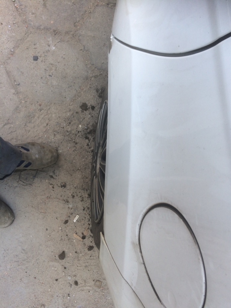 Fragment białego samochodu z widocznym kołem i wlewem paliwa, obok brudny chodnik i fragment buta.