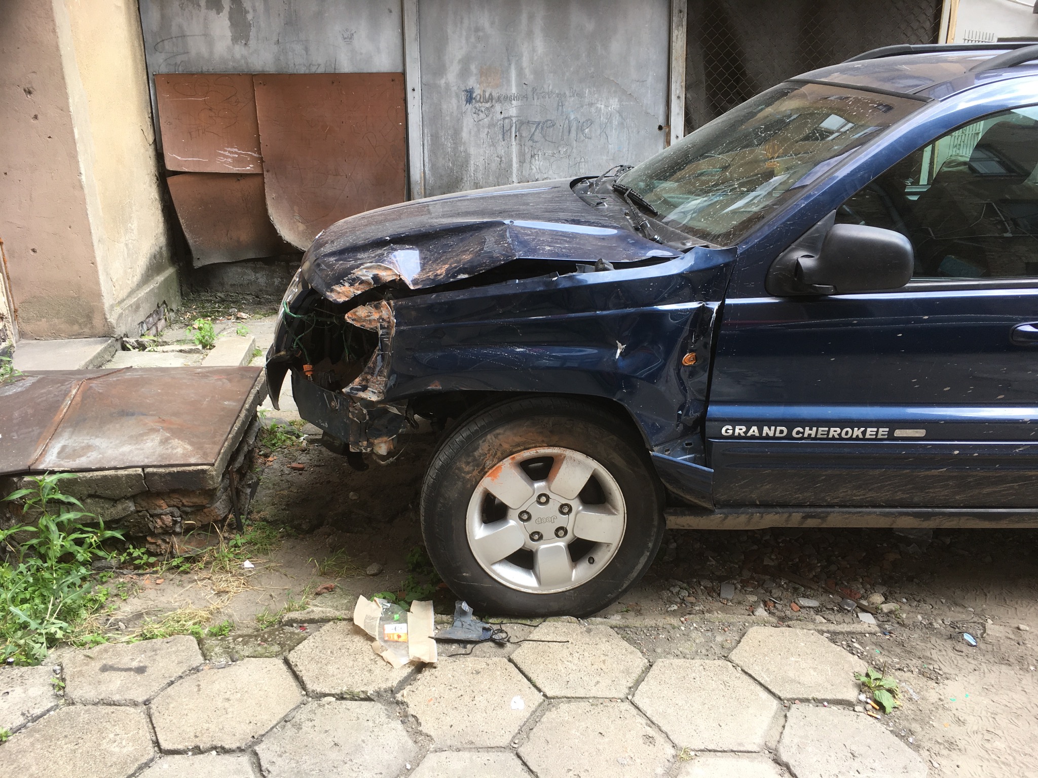 Niebieski Jeep Grand Cherokee po kolizji, z wgniecionym błotnikiem i uszkodzonym przodem, stojący na chodniku z kostki, z widocznymi śladami brudu i uszkodzeń.