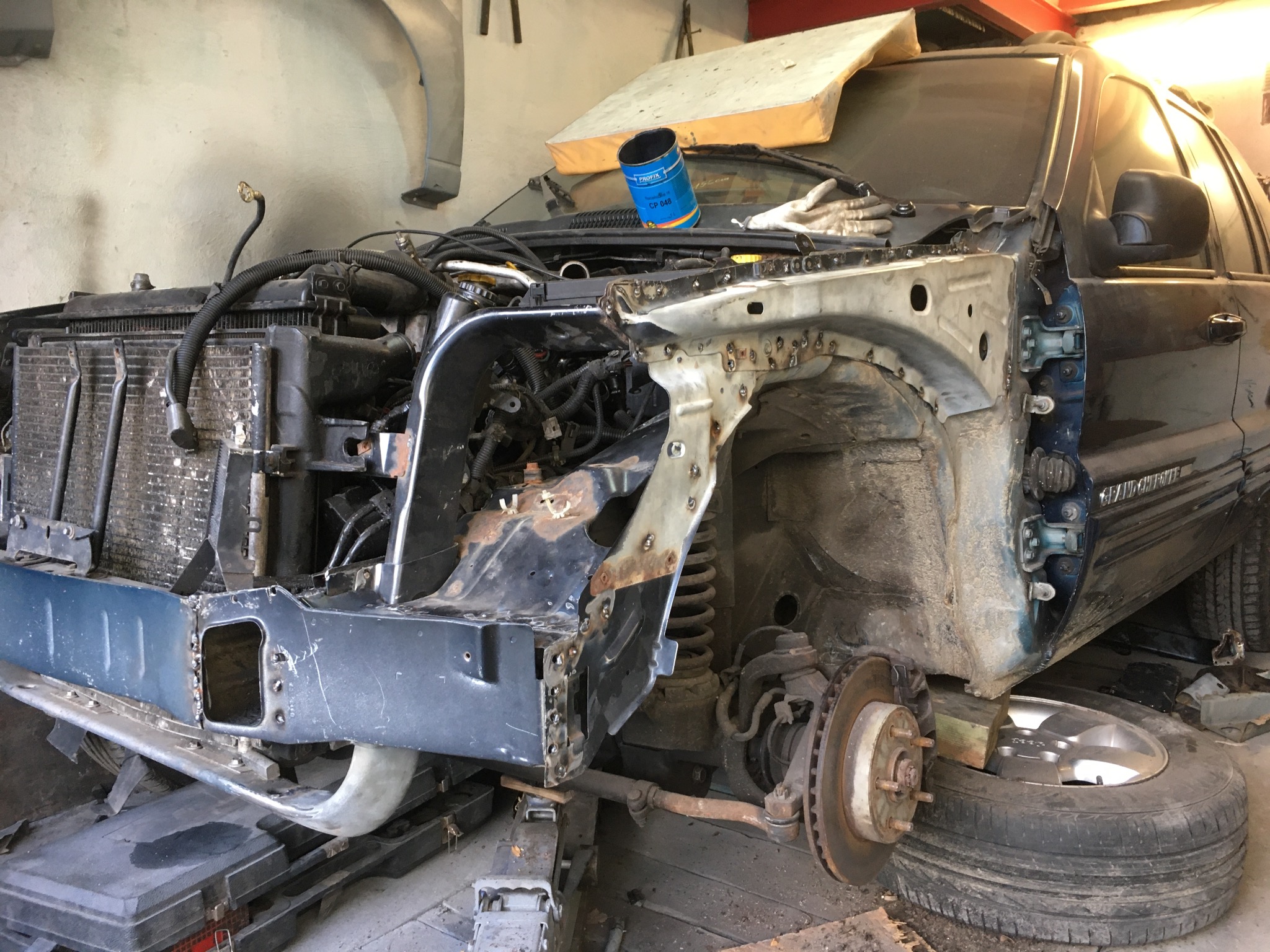 Demontaż i naprawa karoserii samochodu: widoczny fragment nadkola z licznymi otworami po spawaniu, odsłonięty układ zawieszenia i tarcza hamulcowa, puszka z farbą na masce, auto podniesione...