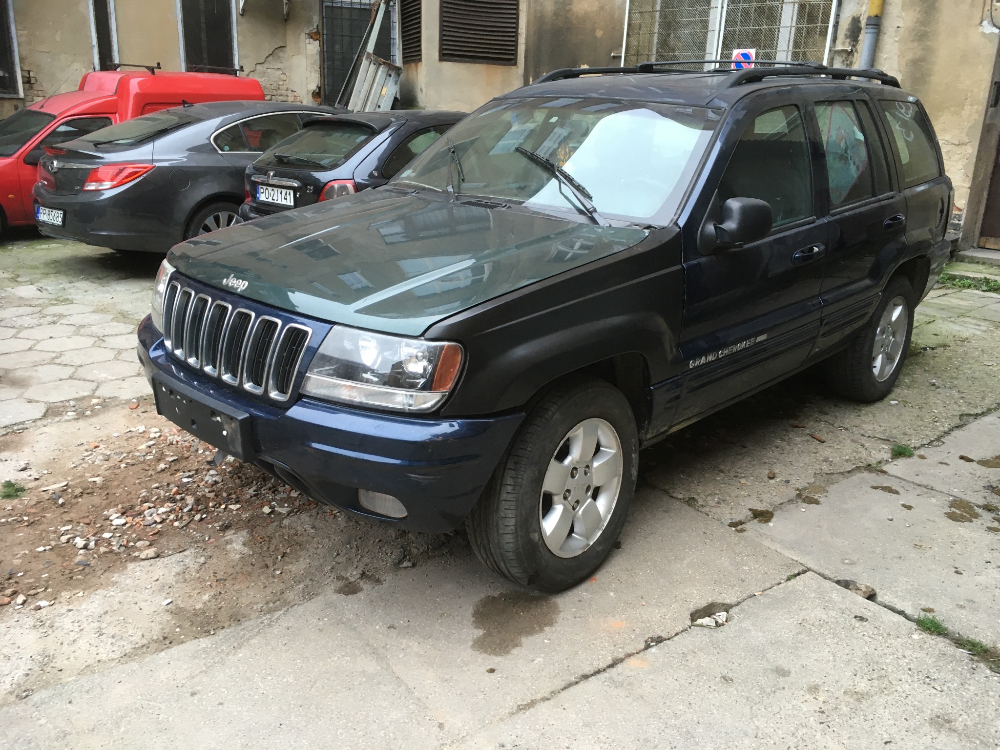 Granatowy Jeep Grand Cherokee z widocznymi śladami korozji na karoserii, zaparkowany na betonowym placu z kilkoma innymi samochodami w tle.