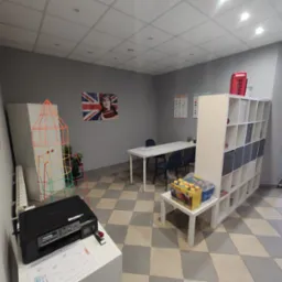 Wnętrze jasnej sali lekcyjnej z biurkami, krzesłami, regałem z zabawkami i dekoracjami, w tym flaga Wielkiej Brytanii na ścianie.