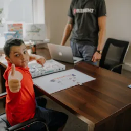 Chłopiec w czerwonej koszulce, siedzący przy biurku, pokazuje kciuk w górę podczas lekcji z nauczycielem, na biurku laptop i materiały edukacyjne.