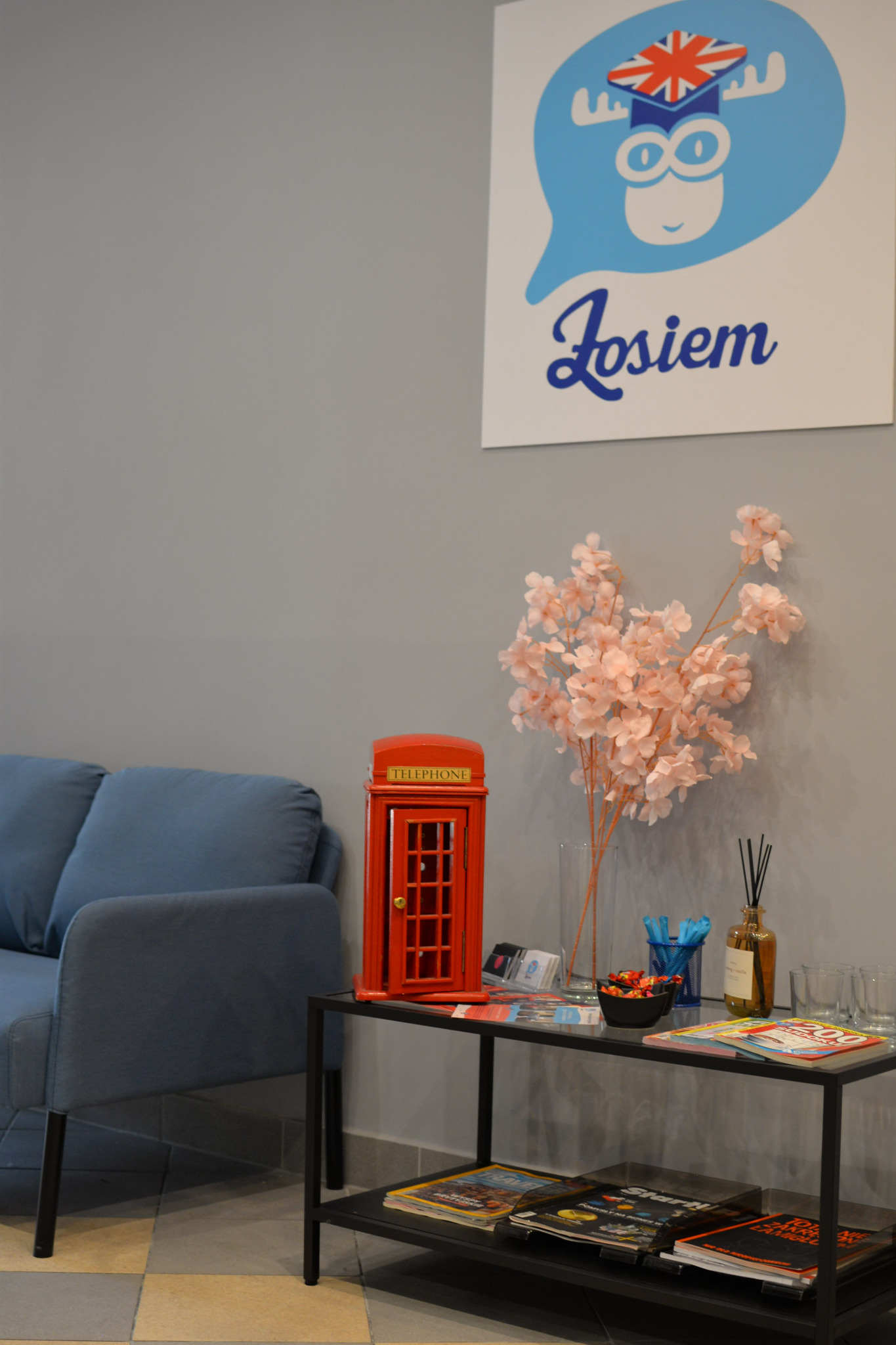 Recepcja szkoły językowej z sofą, stolikiem z dekoracjami: czerwona budka telefoniczna, kwiaty w wazonie, słodycze, ulotki, magazyny i logo szkoły z motywem łosia w birecie z flagą Wielkiej Brytanii.