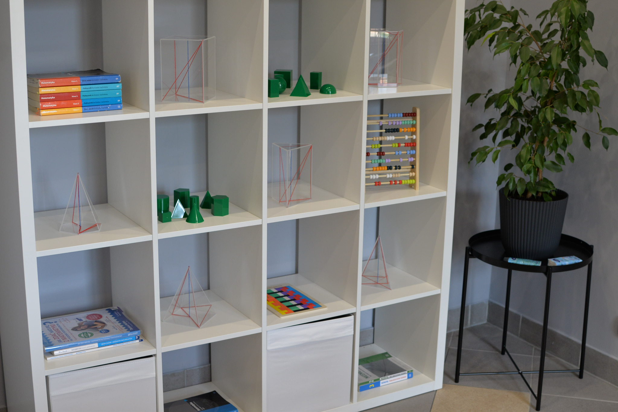 Biały regał z pomocami dydaktycznymi: podręczniki do matematyki, geometryczne bryły z pleksi z kolorowymi krawędziami, drewniane zielone figury geometryczne, kolorowe liczydło. Obok regału stoi...