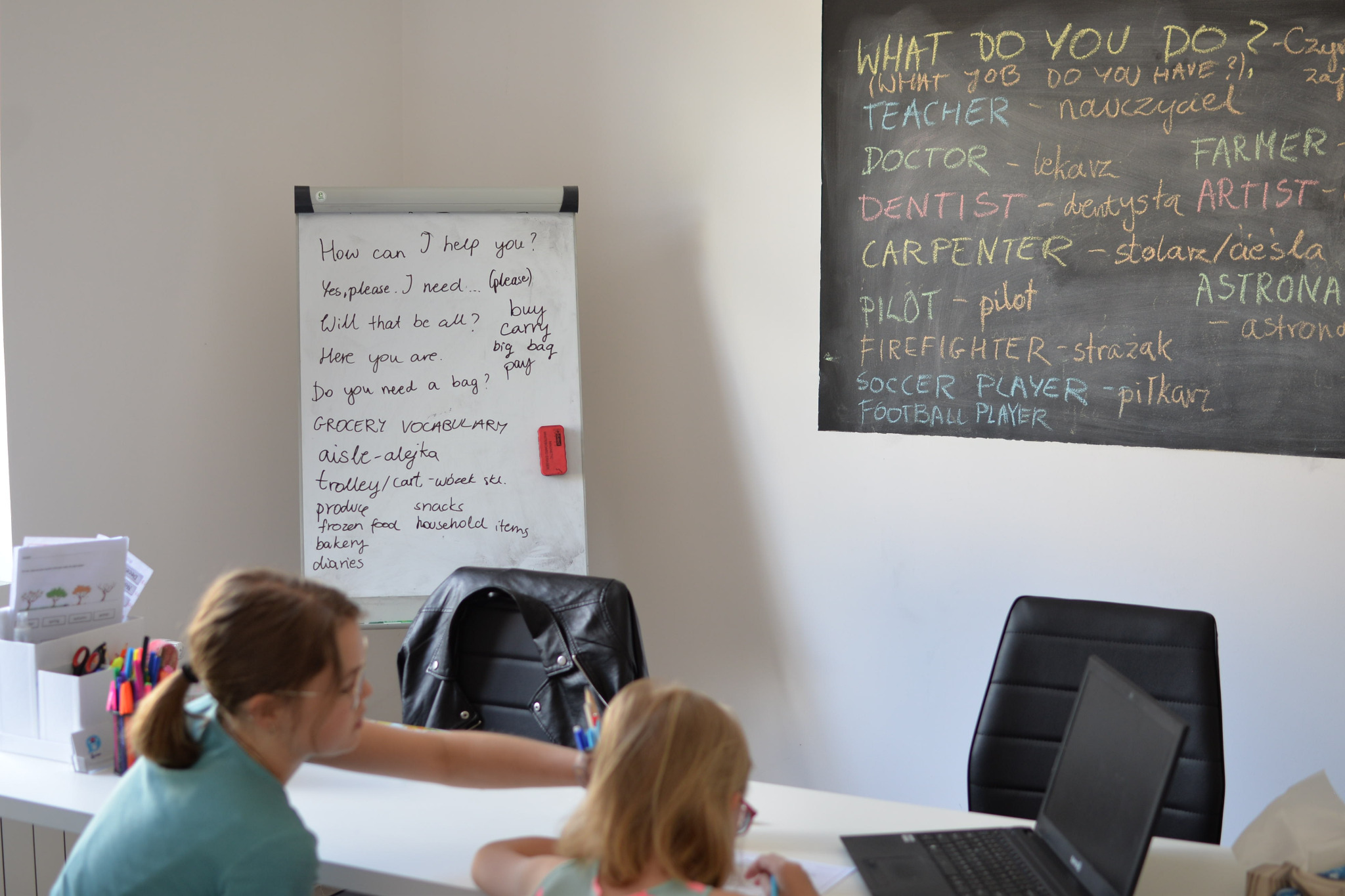Dziewczynka z nauczycielką pracują przy biurku; na tablicy słownictwo związane z zakupami i zawody w języku angielskim.