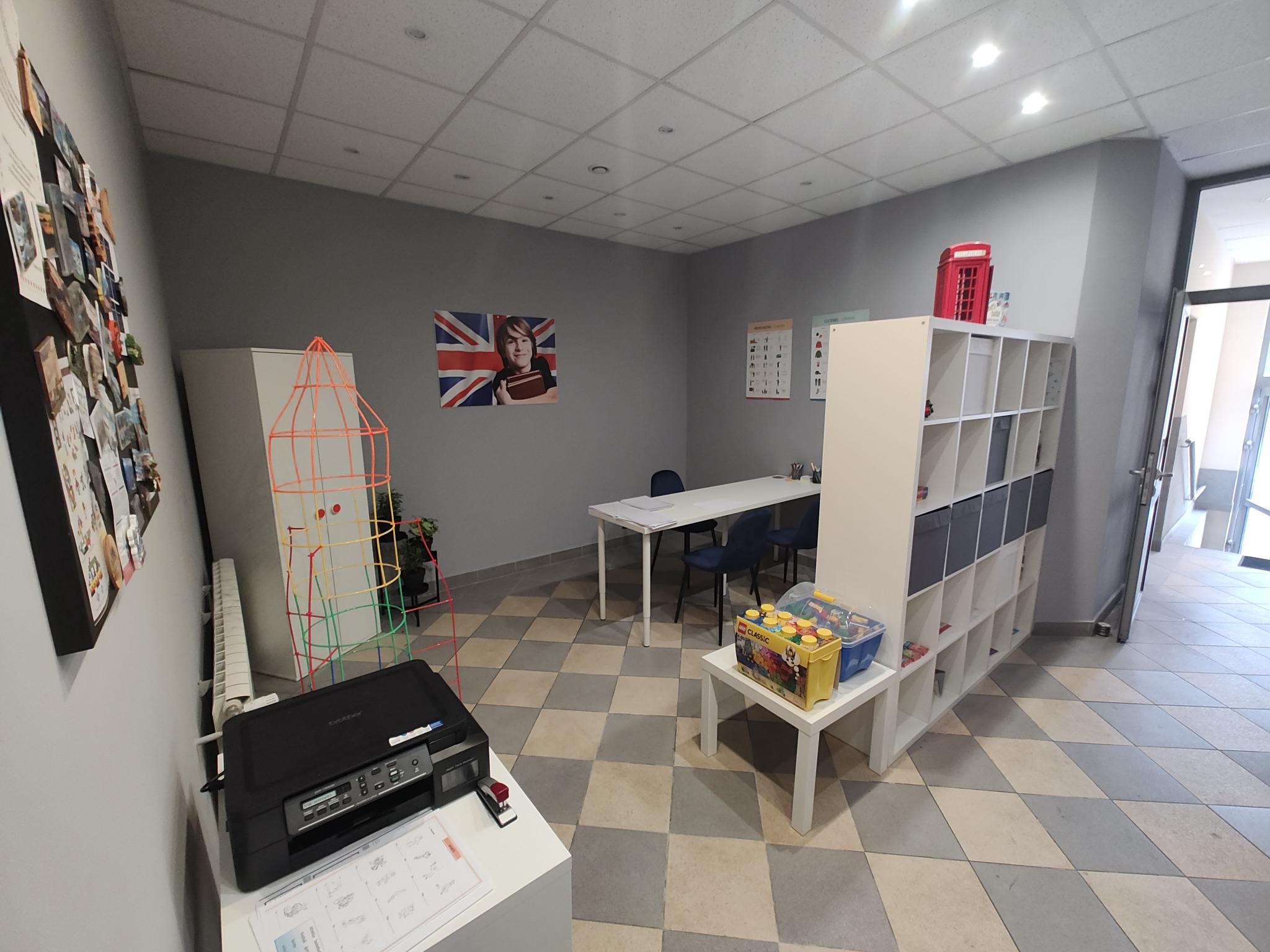 Wnętrze jasnej sali lekcyjnej z biurkami, krzesłami, regałem z zabawkami i dekoracjami, w tym flaga Wielkiej Brytanii na ścianie.