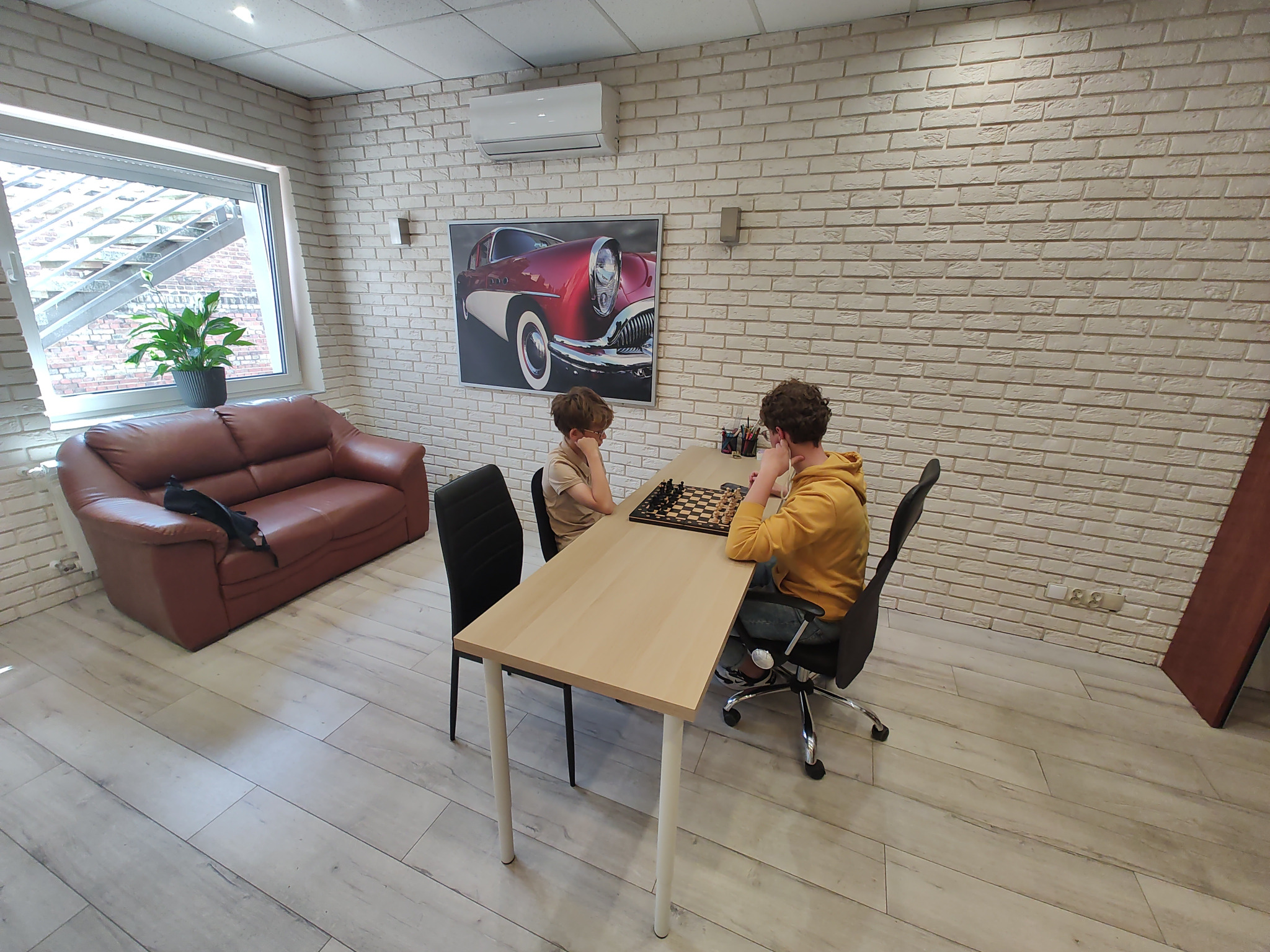 Dwóch chłopców gra w szachy przy jasnym stole w pomieszczeniu z ceglanymi ścianami i obrazem czerwonego samochodu.