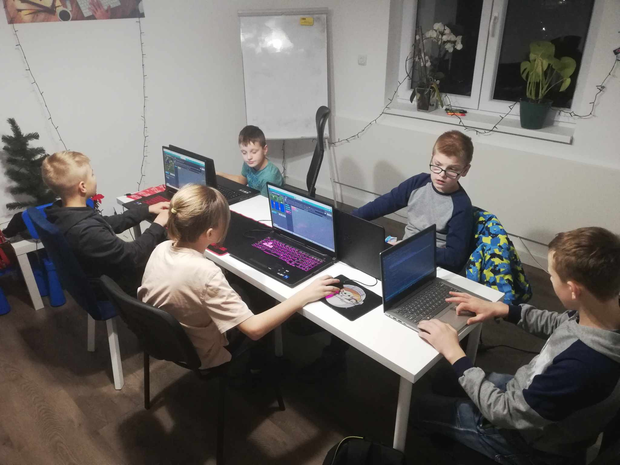 Grupa dzieci siedzi przy białych biurkach z laptopami, ucząc się programowania; na ekranach widoczny kod i gra Minecraft, w tle choinka i dekoracje świąteczne.