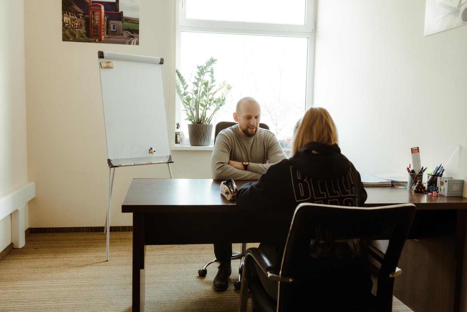 Mężczyzna z brodą siedzi za biurkiem naprzeciwko kobiety w bluzie z kapturem podczas lekcji w jasnym pomieszczeniu z tablicą flipchart i rośliną w doniczce na parapecie.
