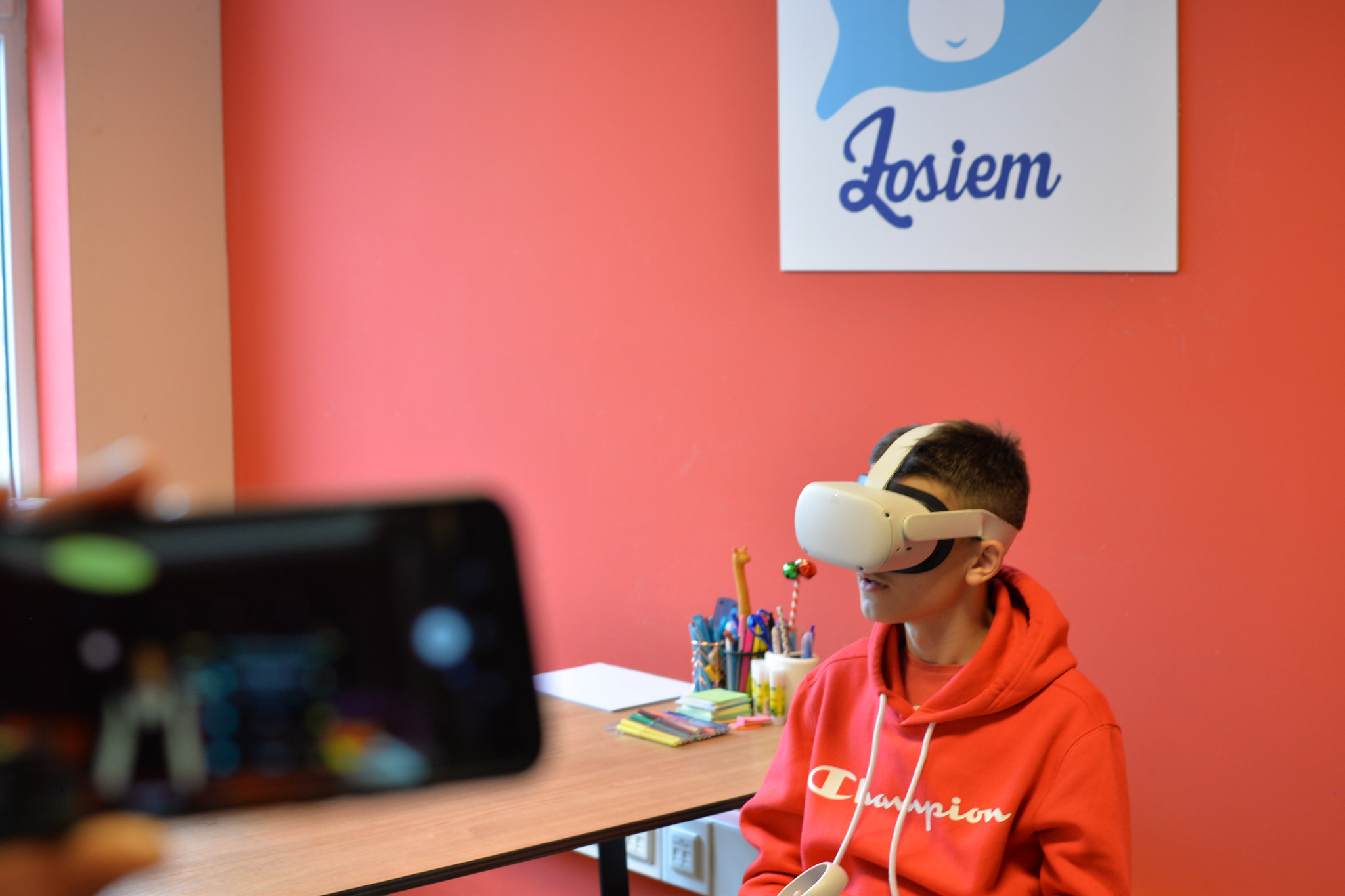 Chłopak w czerwonej bluzie z kapturem i goglach VR siedzi przy biurku, nagrywany telefonem, na tle czerwonej ściany z logo.