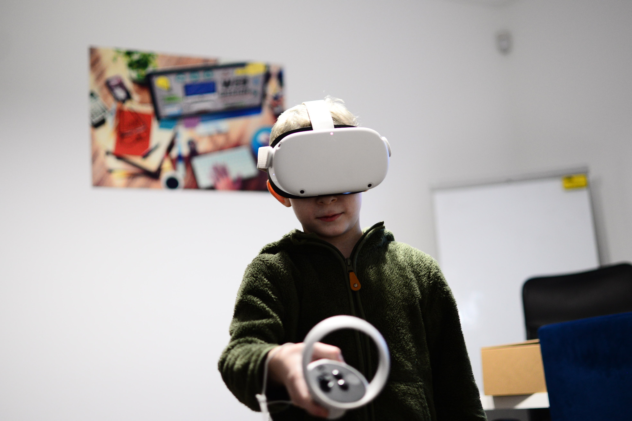 Chłopiec w okularach VR trzymający kontroler, w tle tablica i obrazek na ścianie.