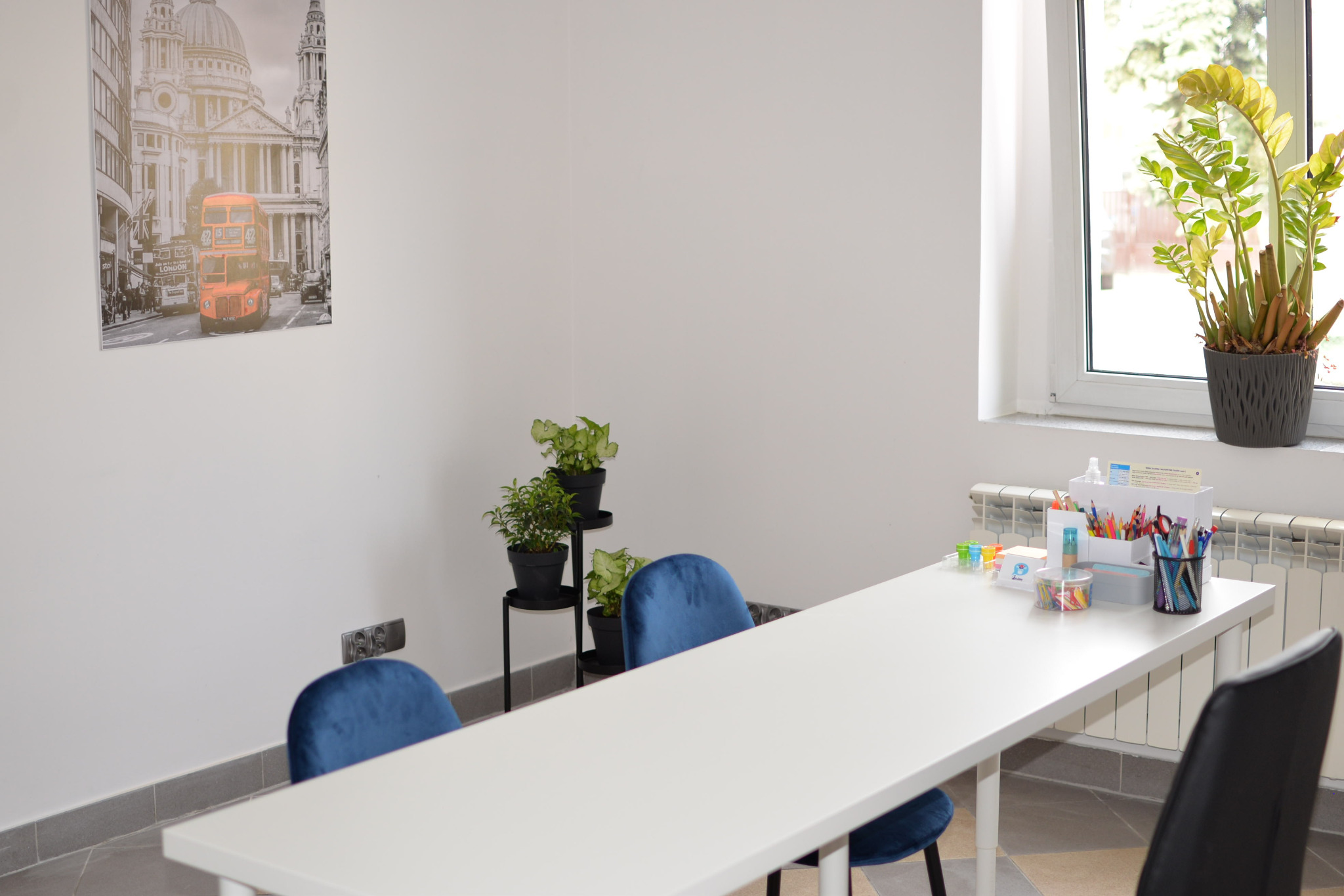 Jasne wnętrze klasy z białym stołem roboczym, niebieskimi krzesłami z aksamitu i roślinami doniczkowymi; na ścianie plakat z londyńskim autobusem i katedrą św. Pawła.
