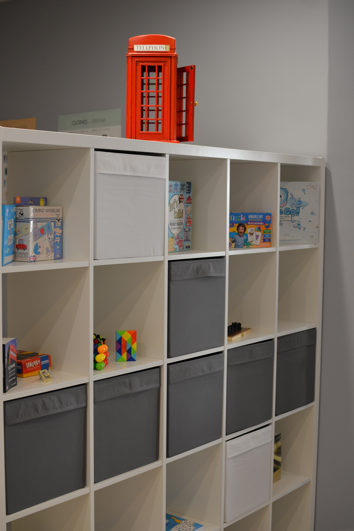 Regał z grami edukacyjnymi, pudełkami i dekoracyjną czerwoną budką telefoniczną, widoczne gry do nauki języka angielskiego i inne gry edukacyjne.