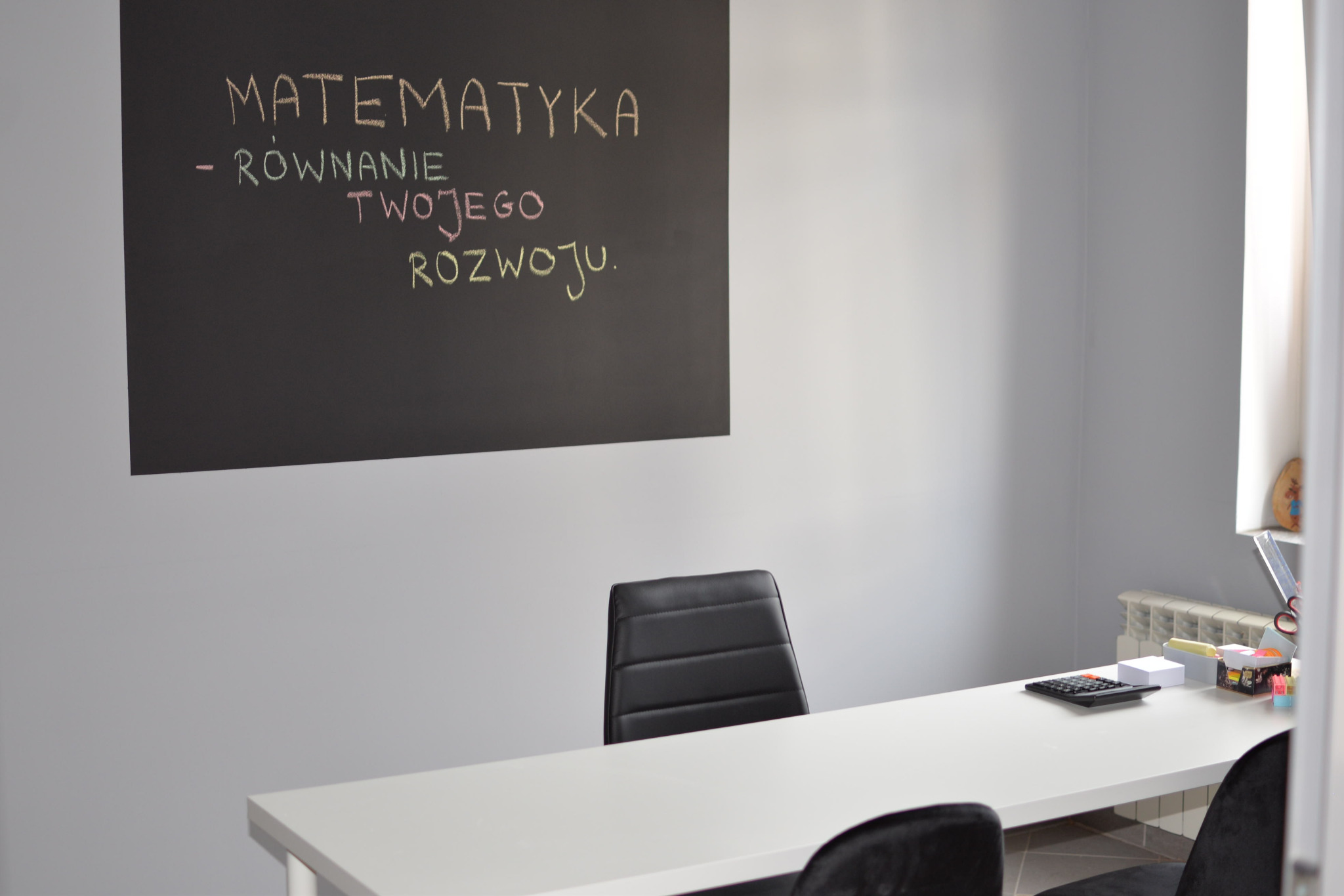 Sala lekcyjna z tablicą z napisem 'Matematyka - równanie Twojego rozwoju', biurkiem, krzesłami i kalkulatorem.