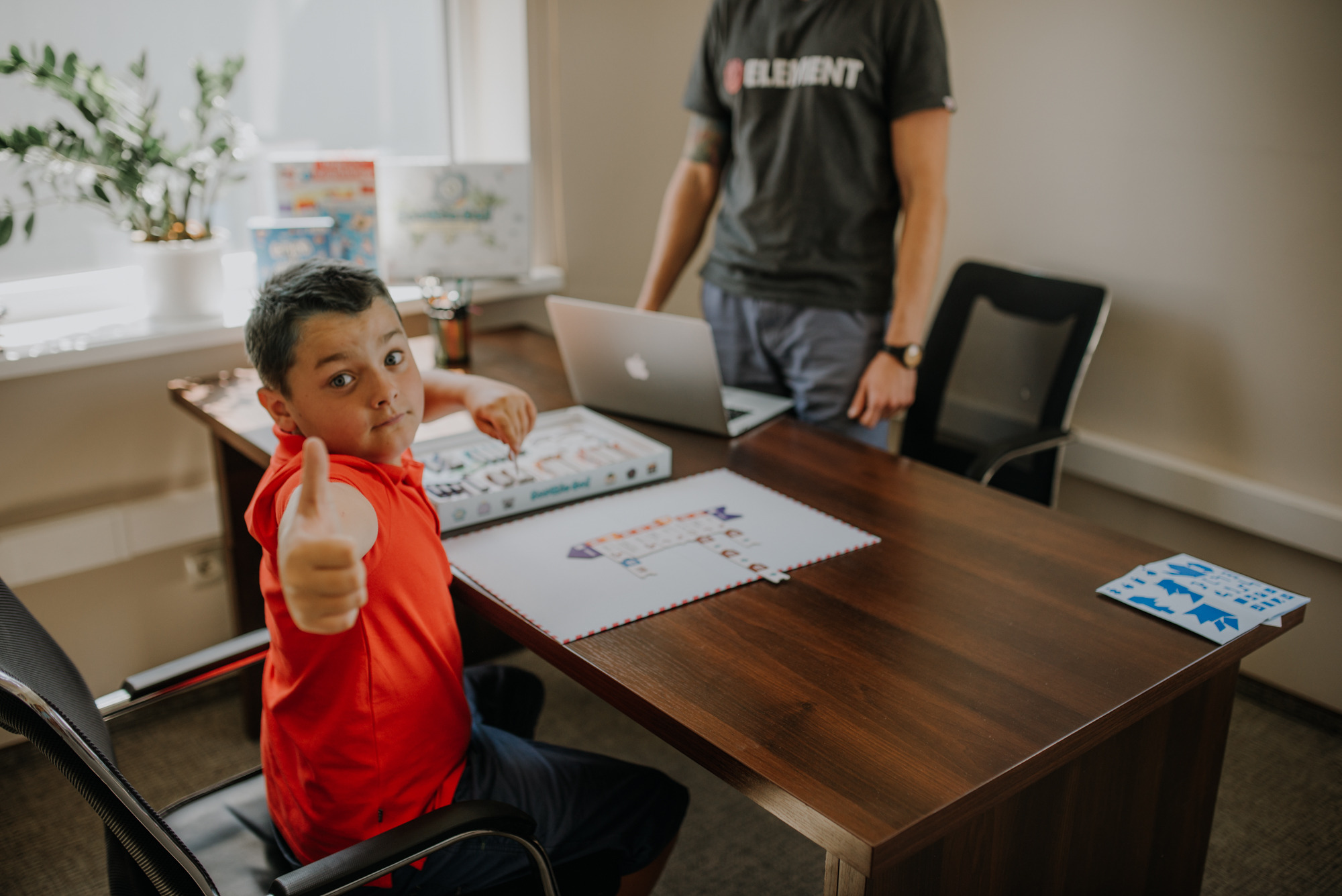 Chłopiec w czerwonej koszulce, siedzący przy biurku, pokazuje kciuk w górę podczas lekcji z nauczycielem, na biurku laptop i materiały edukacyjne.
