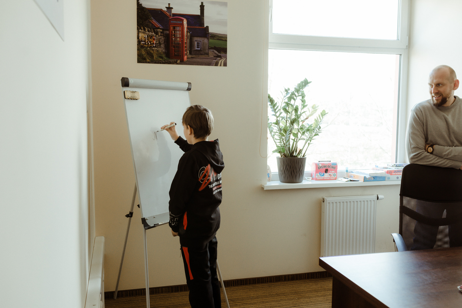Chłopiec pisze markerem na białej tablicy flipchart w jasnym pomieszczeniu z oknem, obok stoi dorosły mężczyzna, na ścianie wisi obrazek z czerwoną budką telefoniczną.