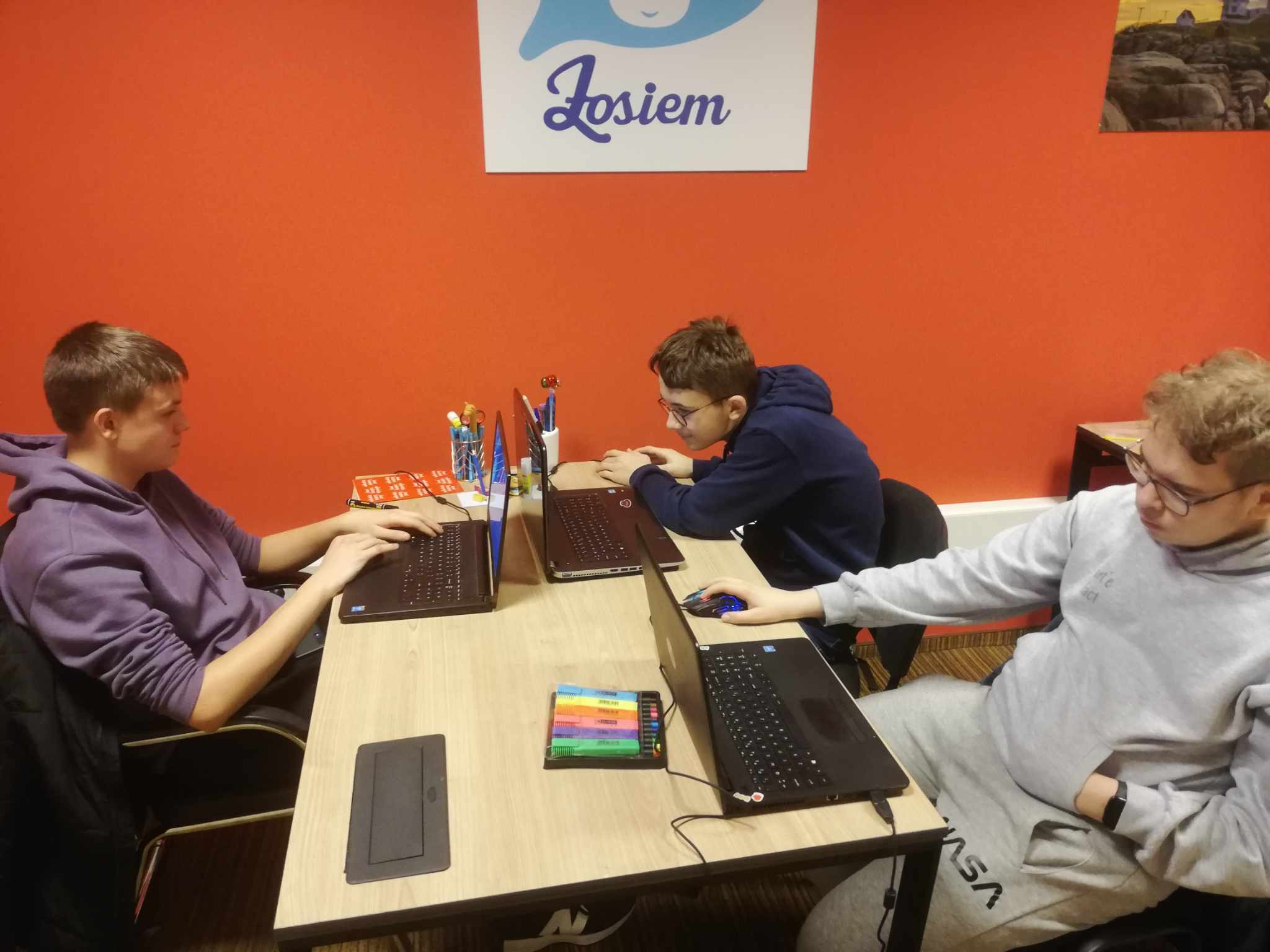 Trzech nastolatków siedzi przy biurku z laptopami, pracując w jasnym pokoju z pomarańczową ścianą i logo 'Josiem' na górze. Na biurku widoczne kolorowe zakreślacze i przybory do pisania.