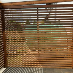 Drewniana pergola z poziomymi listwami, rzucająca cienie na szarą kostkę brukową. W tle widoczne zielone rośliny i fragment białego budynku.