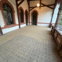 PSLaser Paweł Sklarek - Zadaszony taras z mozaikową posadzką i ceglanymi łukami okiennymi. Drewniana konstrukcja dachu, oświetlenie punktowe, widok na zieleń w tle. Klasyczna architektura.