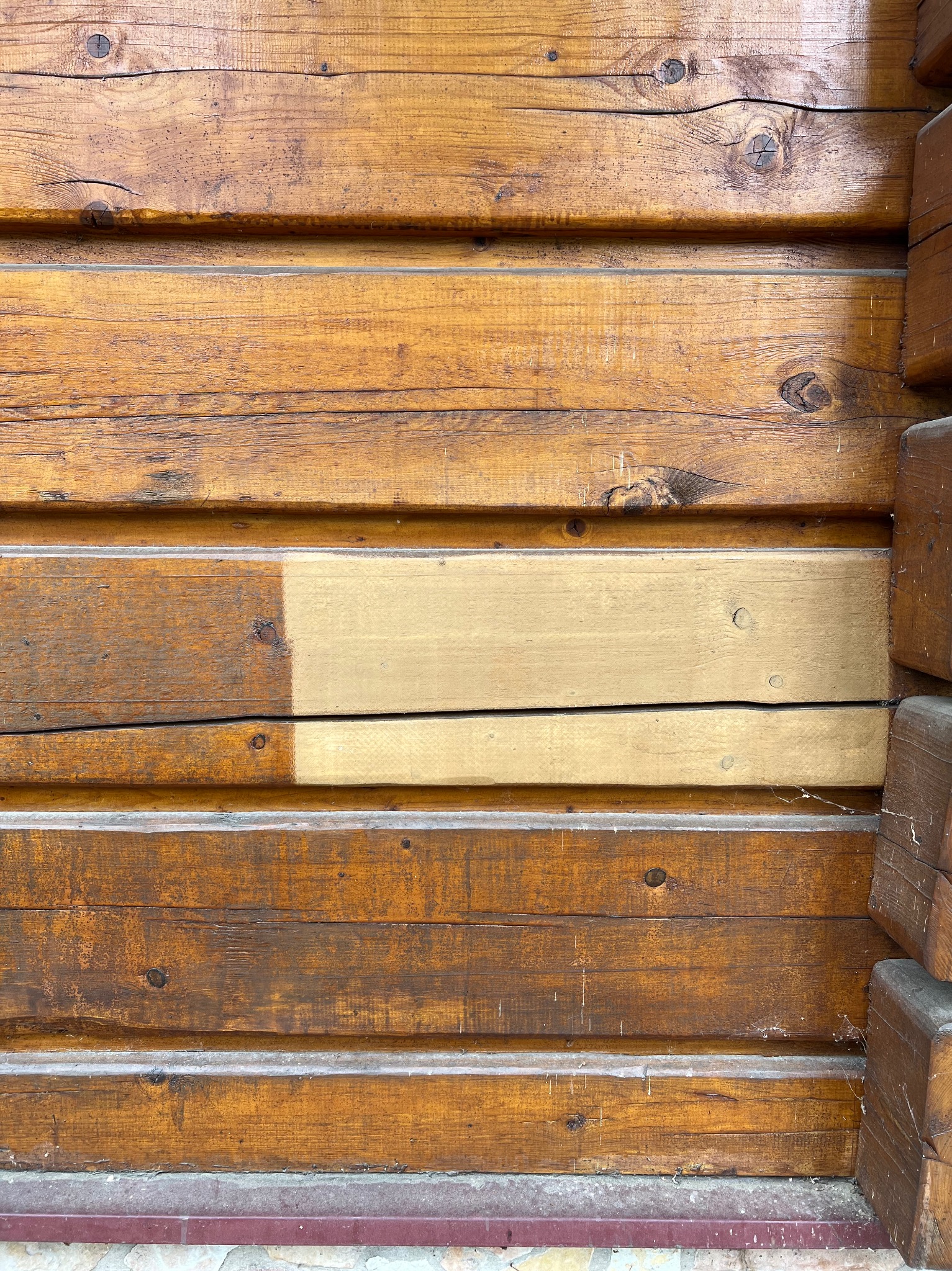 Fragment drewnianej ściany domu z bali, z widocznym obszarem po czyszczeniu lub renowacji, gdzie fragment deski ma jaśniejszy odcień.