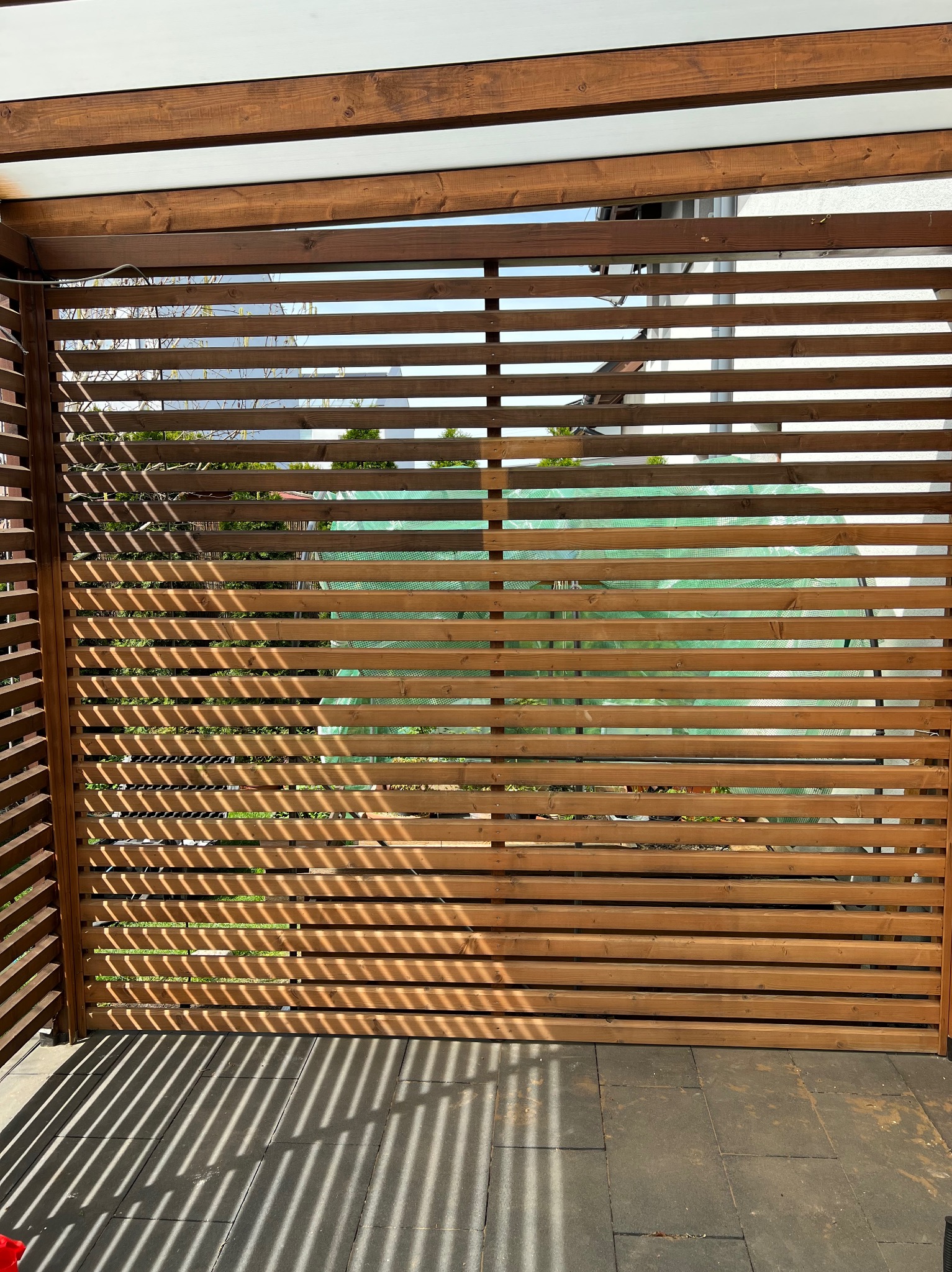 Drewniana pergola z poziomymi listwami, rzucająca cienie na szarą kostkę brukową. W tle widoczne zielone rośliny i fragment białego budynku.