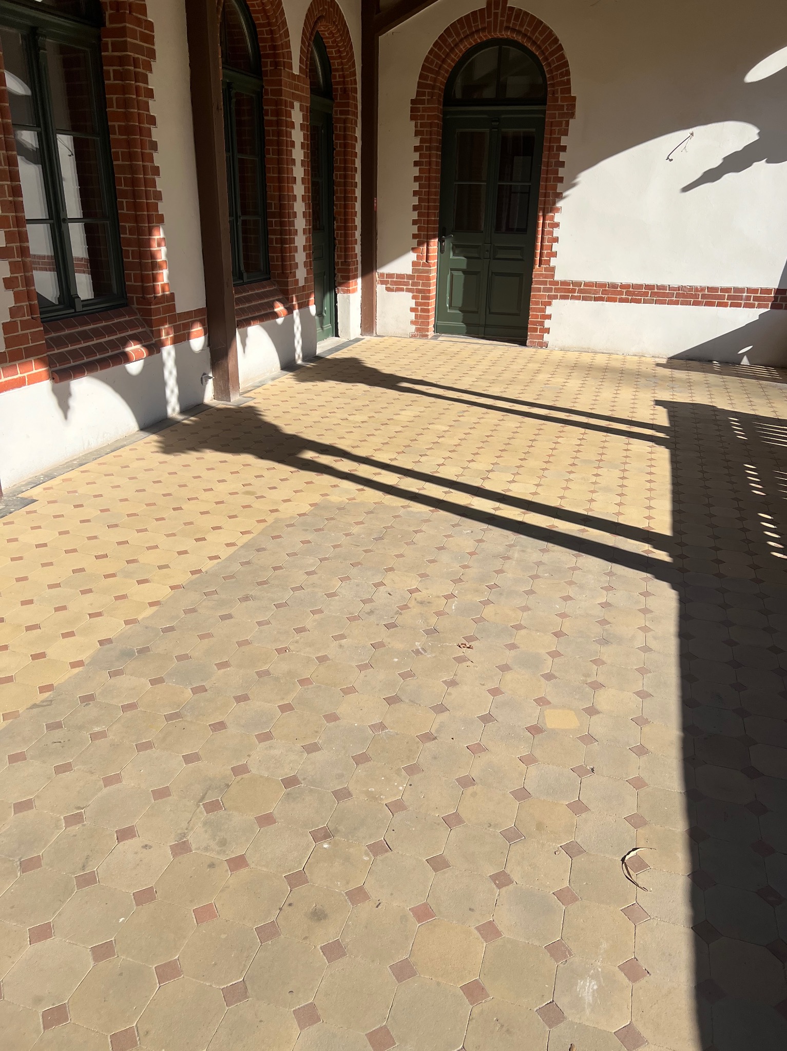 Zewnętrzny dziedziniec z jasną kostką brukową w geometryczny wzór, ceglane łuki okienne i drzwi. Cień rzucany przez konstrukcję dachu.
