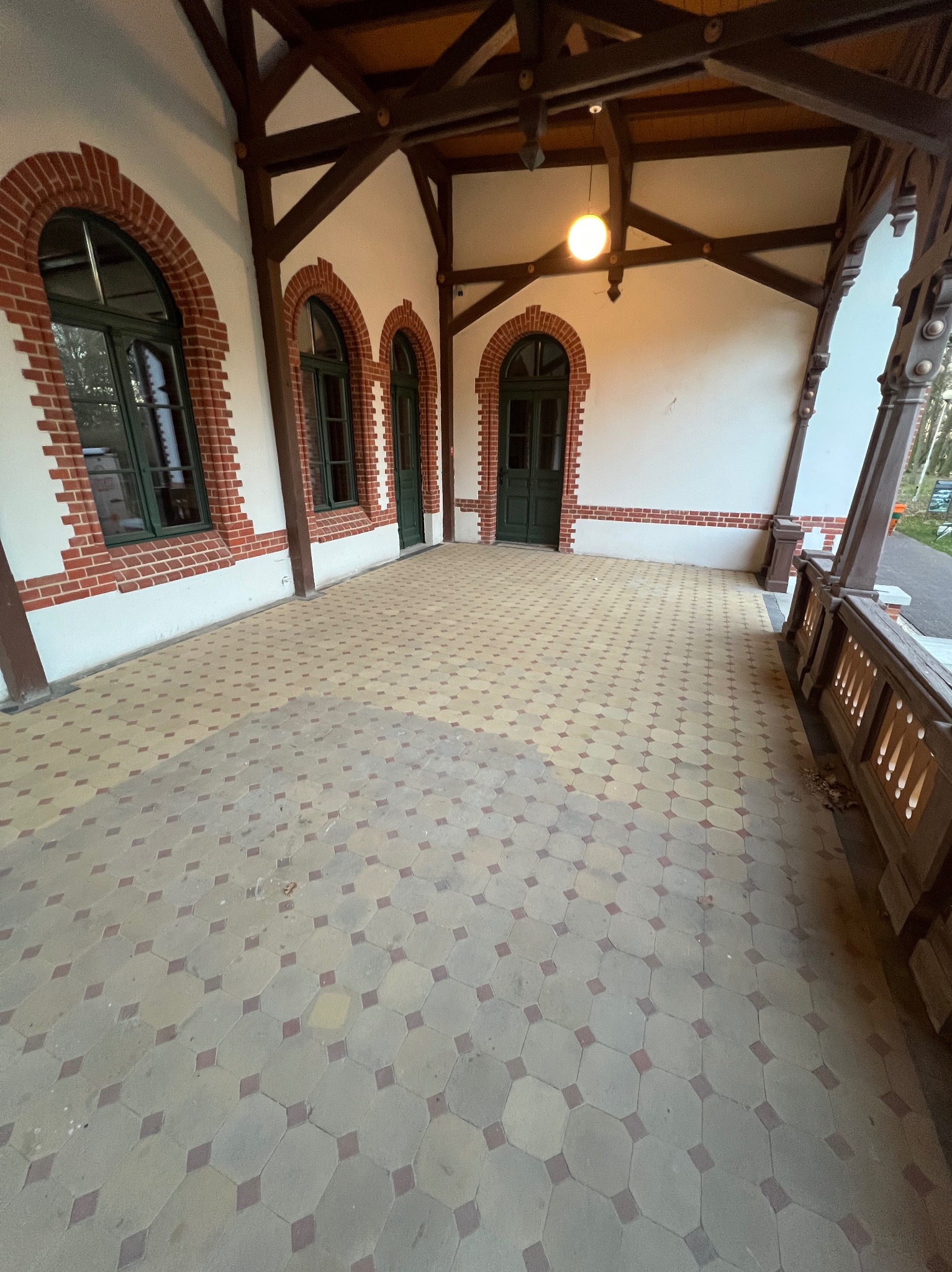 Zadaszony taras z mozaikową posadzką i ceglanymi łukami okiennymi. Drewniana konstrukcja dachu, oświetlenie punktowe, widok na zieleń w tle. Klasyczna architektura.