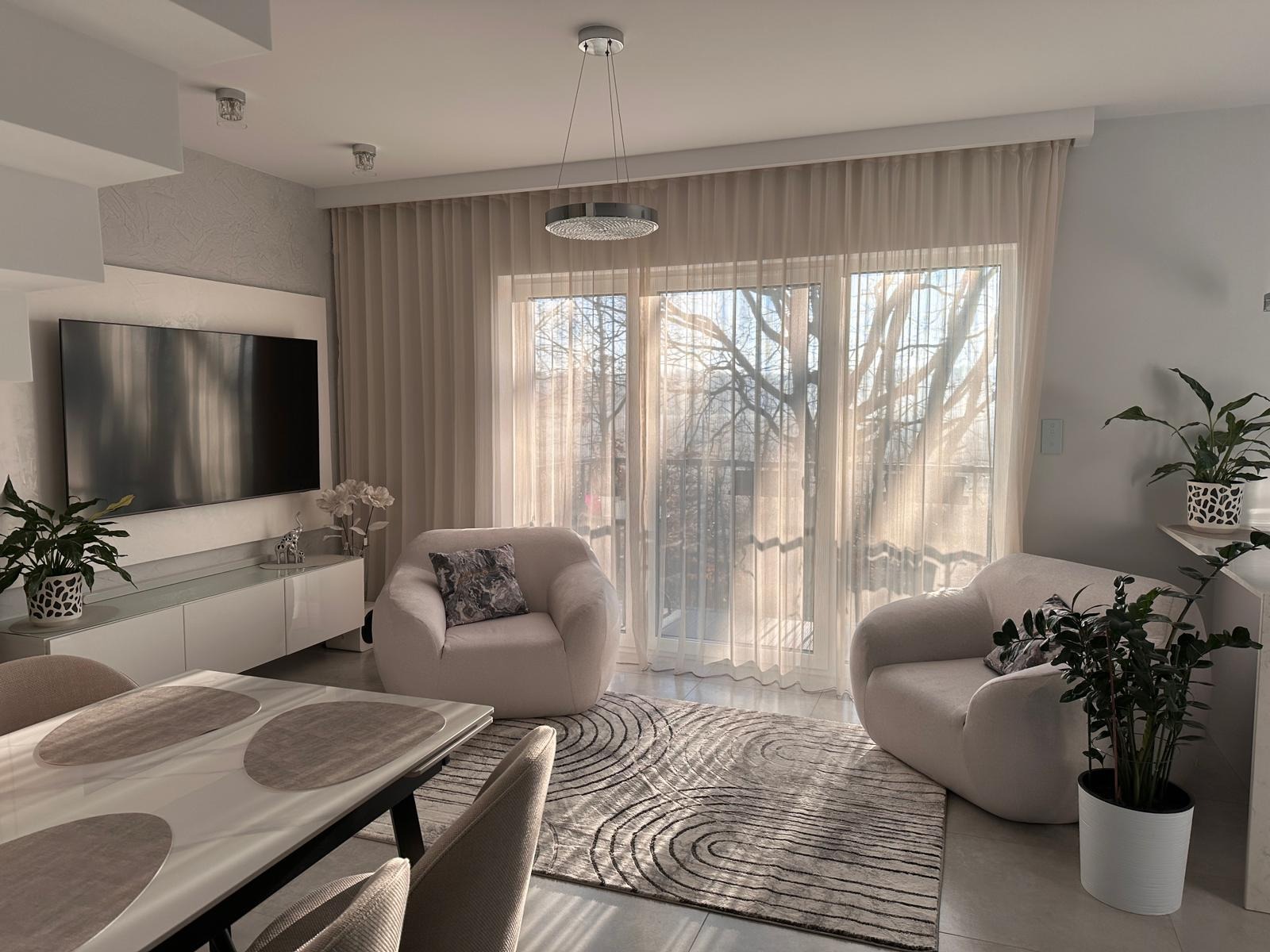 Jasne, przestronne wnętrze salonu z dwoma fotelami typu 'bear', dużym oknem z widokiem na drzewa, dywanem w geometryczne wzory oraz nowoczesnym oświetleniem sufitowym.