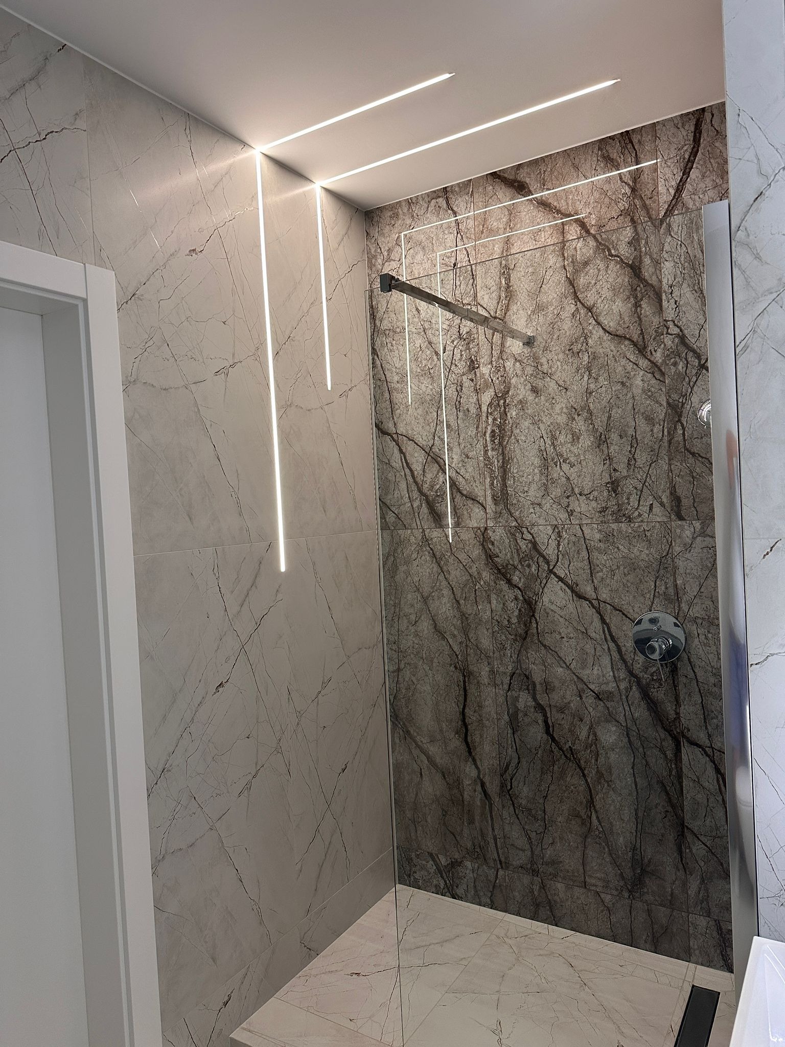 Nowoczesna kabina prysznicowa z marmurowymi płytkami w odcieniach szarości i brązu, oświetlona liniowymi lampami LED w suficie i na ścianach.