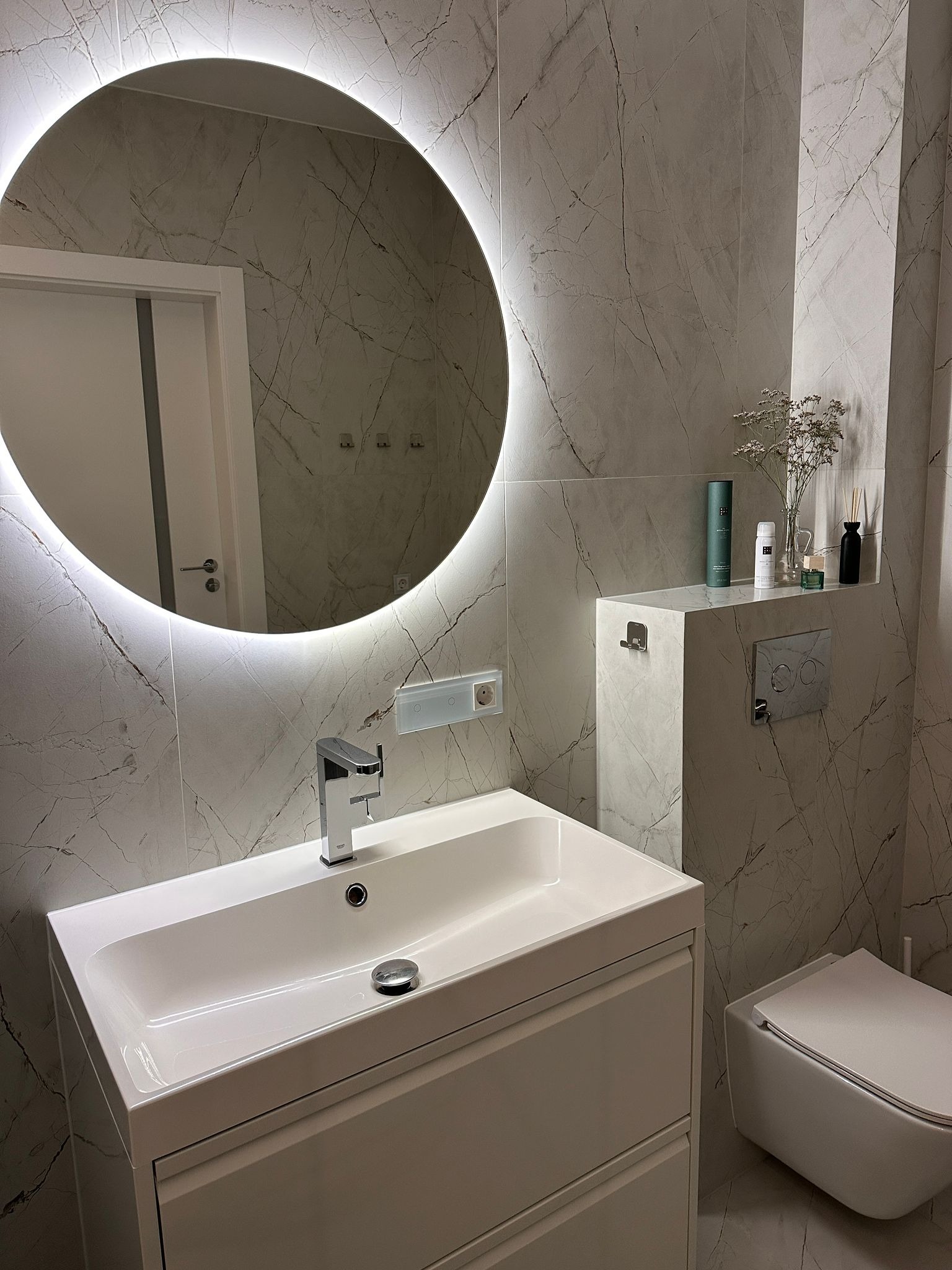 Nowoczesna łazienka z umywalką nablatową, podświetlanym okrągłym lustrem, marmurowymi płytkami ściennymi i wiszącą toaletą.