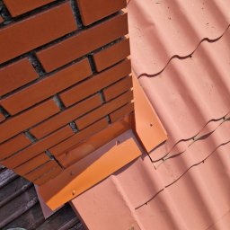 Piotr kowalski - Czerwona dachówka ceramiczna z obróbką blacharską komina, widoczne mocowania i fragment buta osoby wykonującej prace.