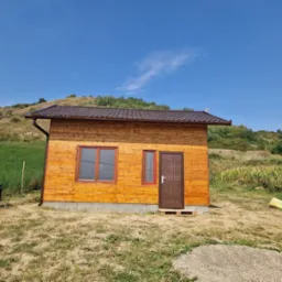 Drewniany domek z brązowym dachem i rynnami, otoczony zielenią wzgórz i błękitnym niebem.