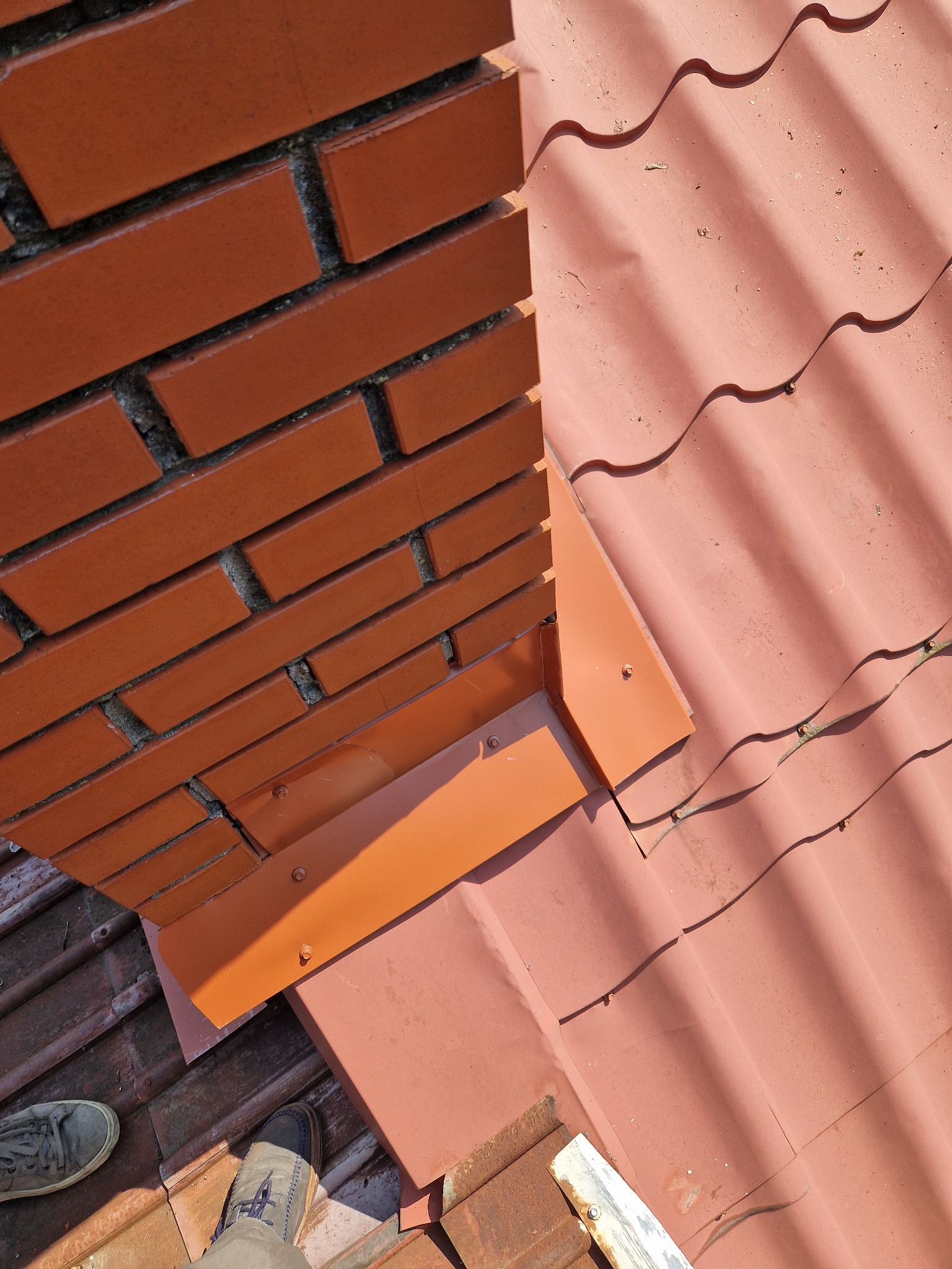Czerwona dachówka ceramiczna z obróbką blacharską komina, widoczne mocowania i fragment buta osoby wykonującej prace.