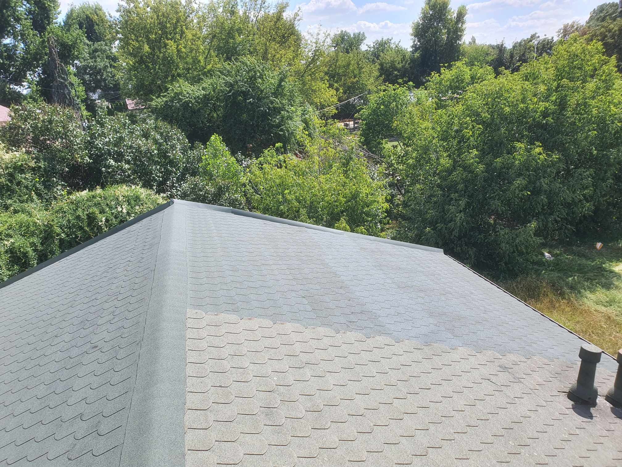 Widok z góry na dach pokryty szarym gontem bitumicznym, z widocznym fragmentem świeżo położonego pasa gontu wzdłuż kalenicy, otoczony zielenią drzew.