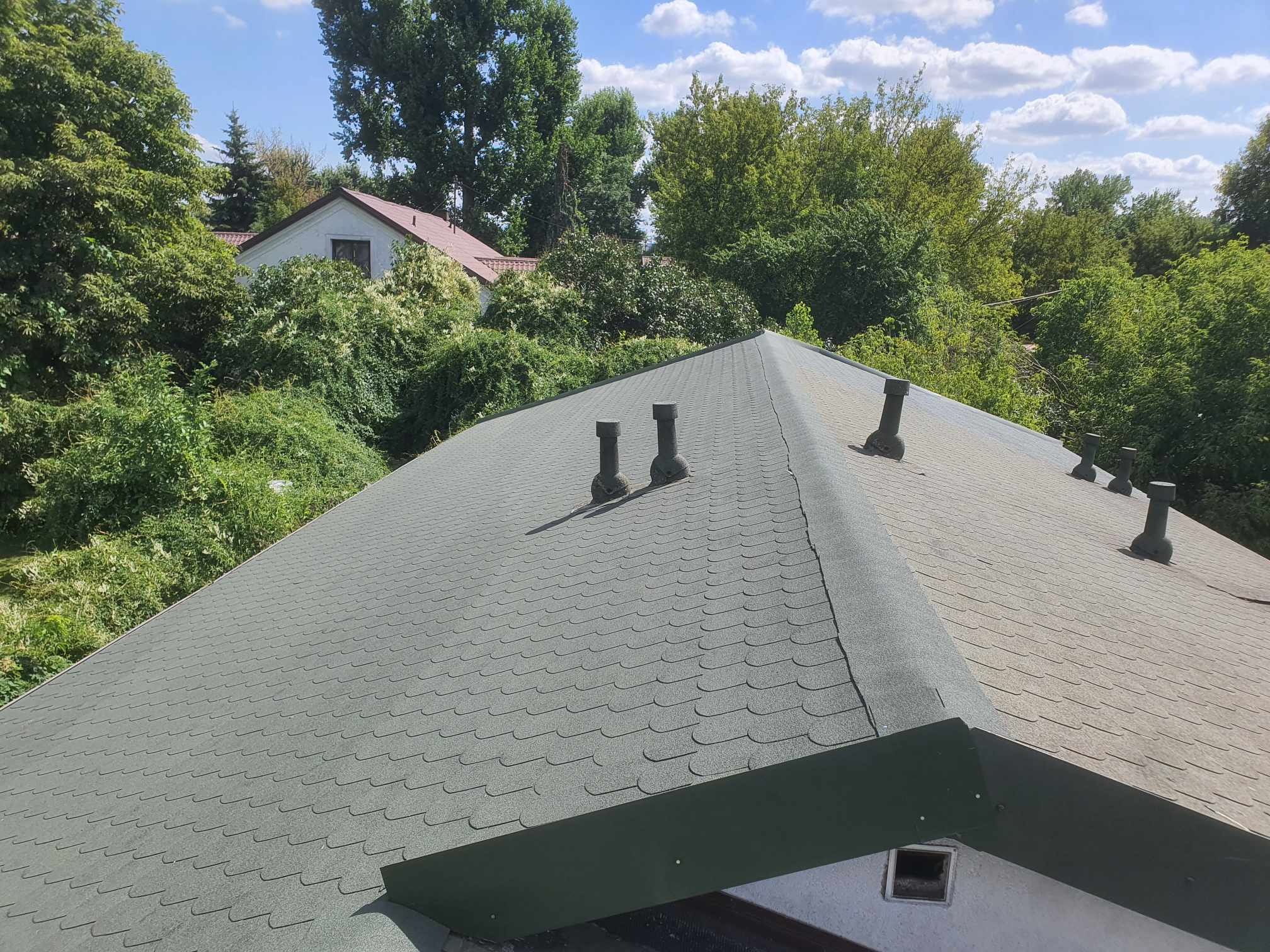 Widok z góry na nowy dach pokryty gontem bitumicznym w odcieniach szarości, z widocznymi kominkami wentylacyjnymi i obróbkami blacharskimi w kolorze ciemnozielonym, na tle bujnej zieleni drzew...