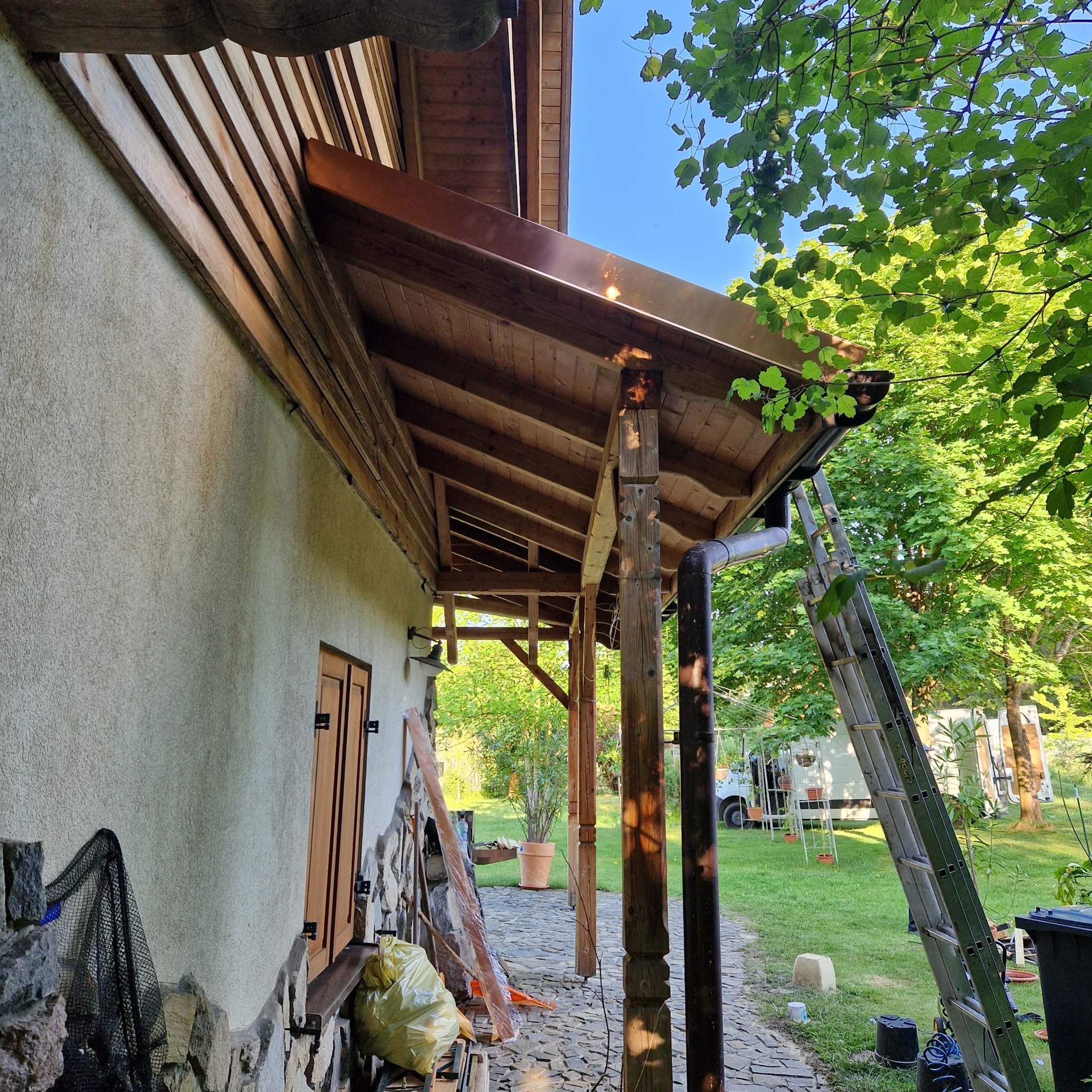 Dom z drewnianym podbitką i miedzianymi rynnami, widoczna drabina oparta o rynnę spustową, w tle zieleń ogrodu i zaparkowany samochód dostawczy.
