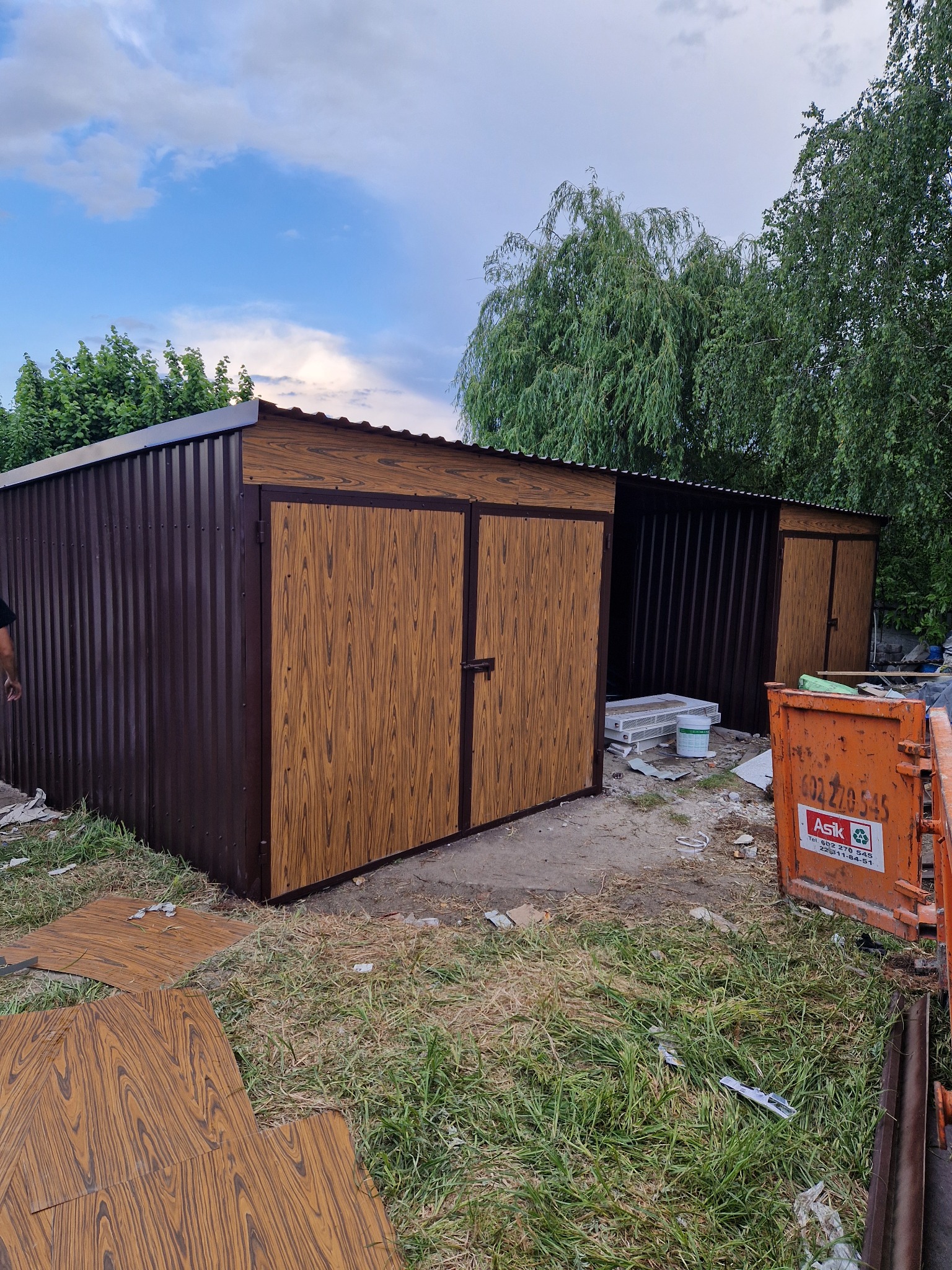 Montaż brązowego garażu blaszanego z drzwiami imitującymi drewno, widoczny kontener na gruz i elementy wykończeniowe na trawie.