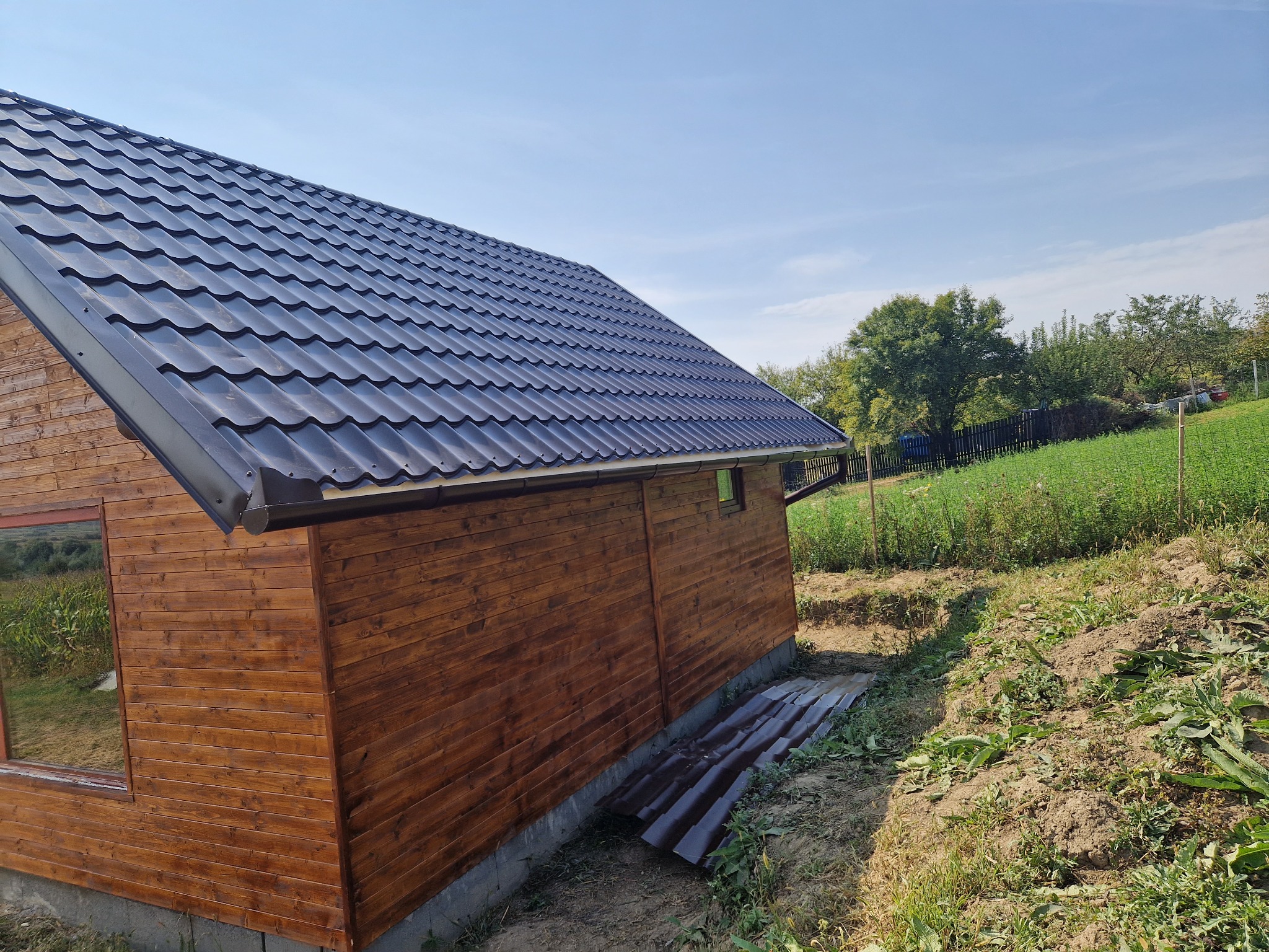 Nowoczesny, ciemny dach z blachy trapezowej na drewnianym domku, z widoczną rynną i otoczeniem działki z zielenią.