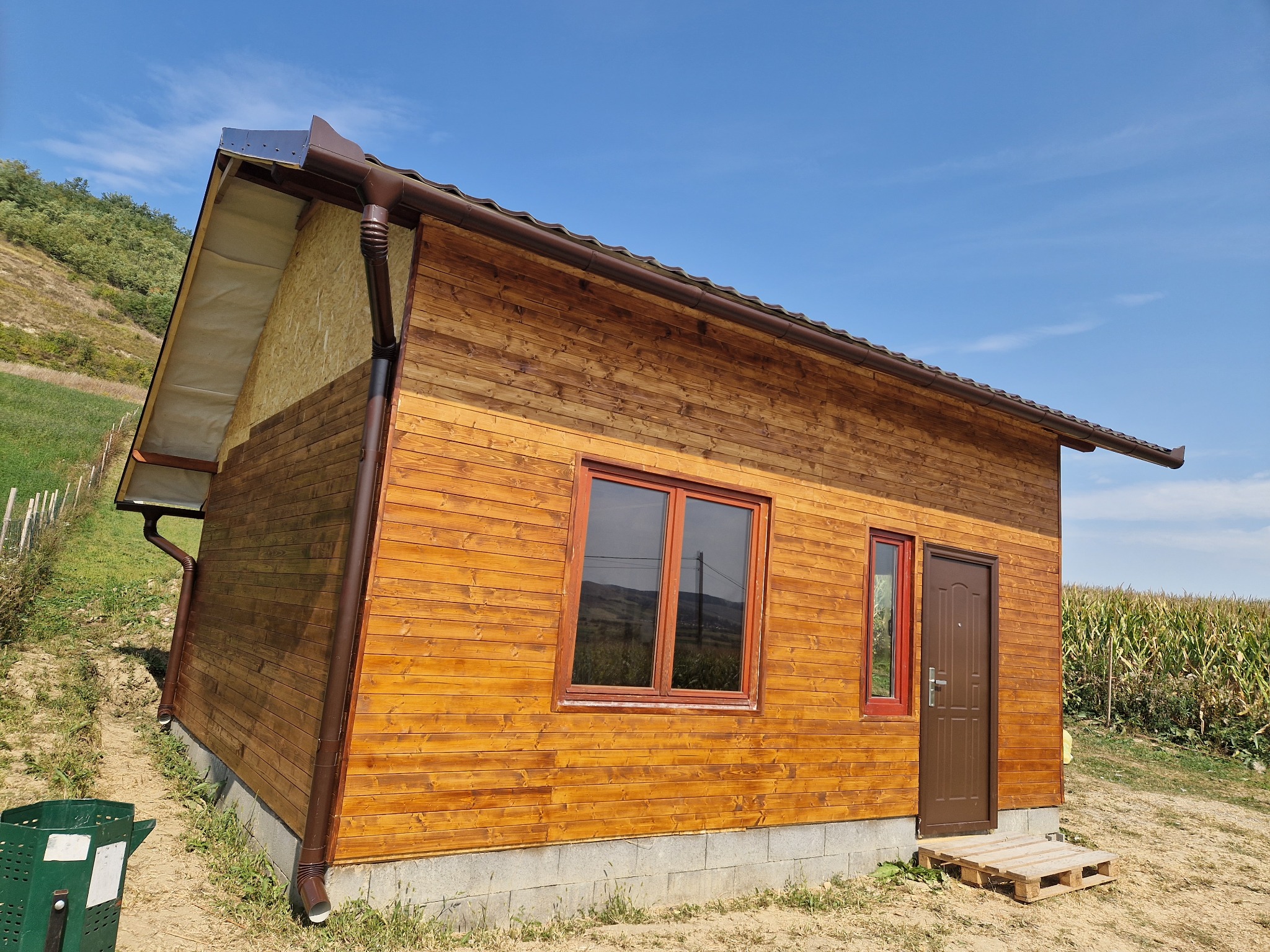 Nowo wybudowany drewniany dom z brązowymi rynnami i obróbkami blacharskimi, widok z zewnątrz na tle błękitnego nieba i zieleni.