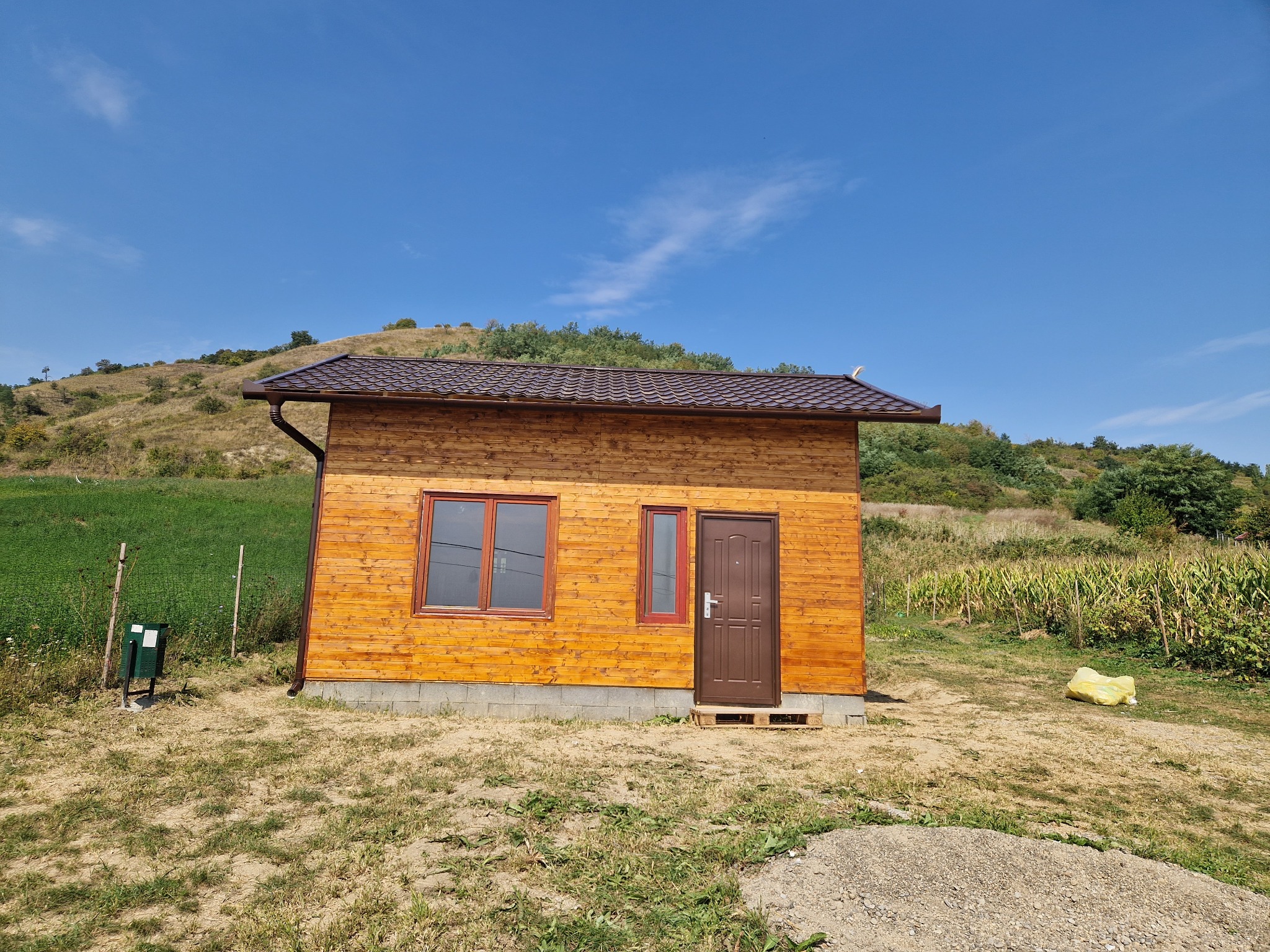 Drewniany domek z brązowym dachem i rynnami, otoczony zielenią wzgórz i błękitnym niebem.