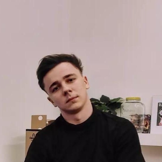 Portret młodego mężczyzny w czarnej koszulce na tle białej ściany z dekoracjami na półce, w tym roślina doniczkowa i słoik z żółtą pokrywką.