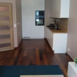 Nowoczesne wnętrze mieszkania z drewnianą podłogą, białą zabudową kuchenną i jasną, drewnianą stolarką drzwiową.