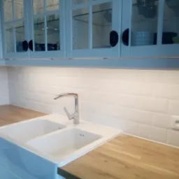 Biała, dwukomorowa ceramiczna umywalka pod oknem w kuchni z białymi, przeszklonymi szafkami i drewnianym blatem, oświetlona lampą podszafkową.