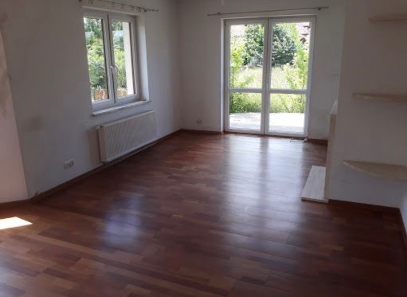 Pusty pokój z nowo ułożoną, ciemną podłogą z paneli, widok na ogród przez duże okno balkonowe z białymi ramami, białe ściany i grzejnik pod oknem.