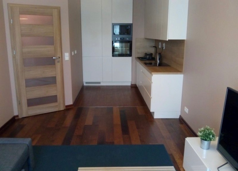 Nowoczesne wnętrze mieszkania z drewnianą podłogą, białą zabudową kuchenną i jasną, drewnianą stolarką drzwiową.