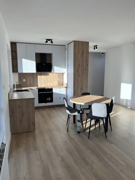 Nowoczesna kuchnia z białymi szafkami i drewnianymi akcentami, okrągły stół z krzesłami w tle, panele podłogowe w odcieniach drewna, oświetlenie punktowe na suficie.