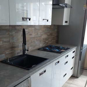 Błyszcząca, nowoczesna kuchnia z czarnym, głębokim zlewem i wysoką baterią, szarym blatem, białymi szafkami z poziomymi uchwytami, okładziną ściany imitującą kamień i płytą gazową zabezpieczoną folią.