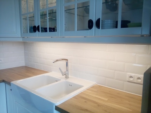 Biała, dwukomorowa ceramiczna umywalka pod oknem w kuchni z białymi, przeszklonymi szafkami i drewnianym blatem, oświetlona lampą podszafkową.